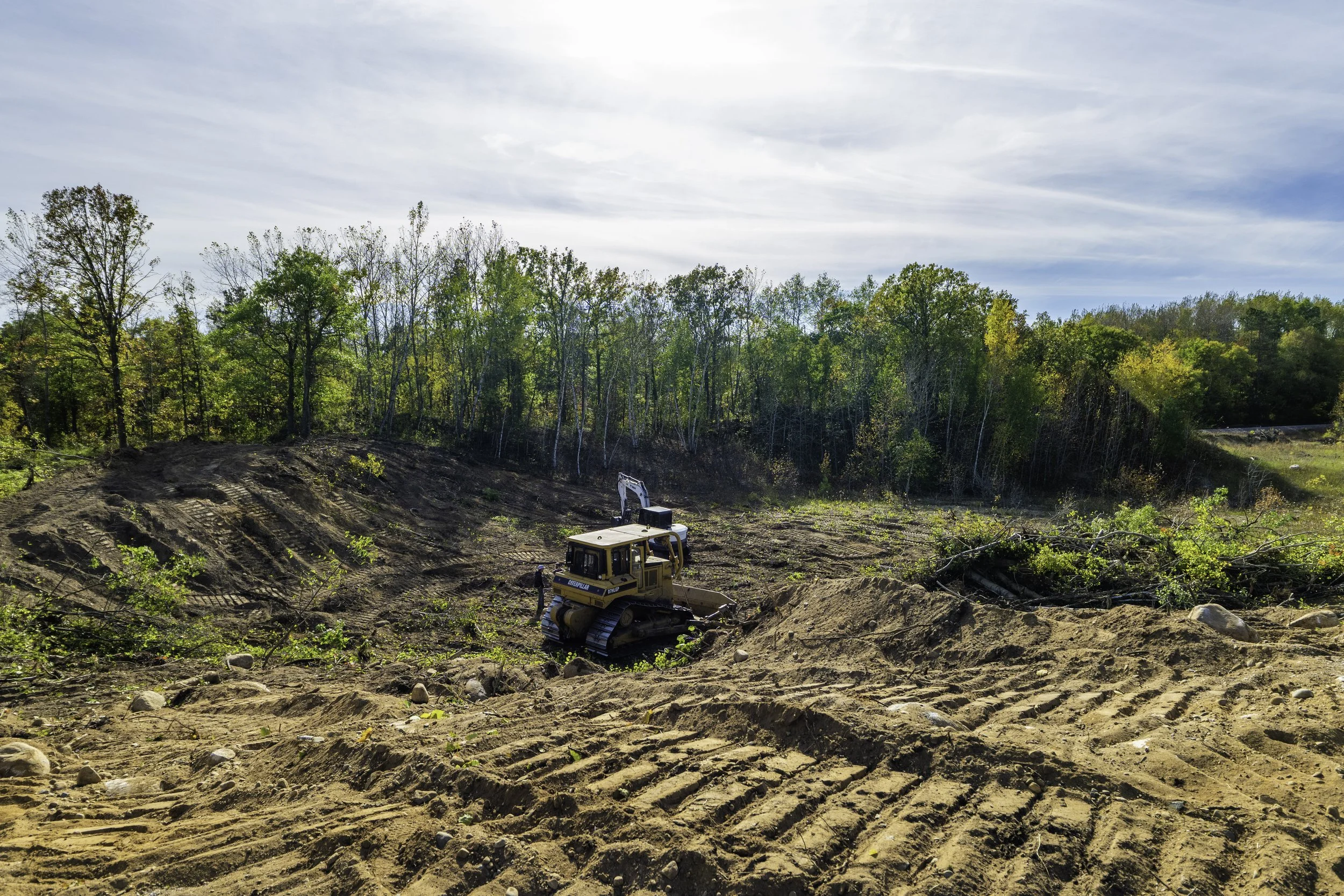 Construction site with bulldozer and excavator clearing land, surrounded by trees with changing leaves under a partly cloudy sky.
