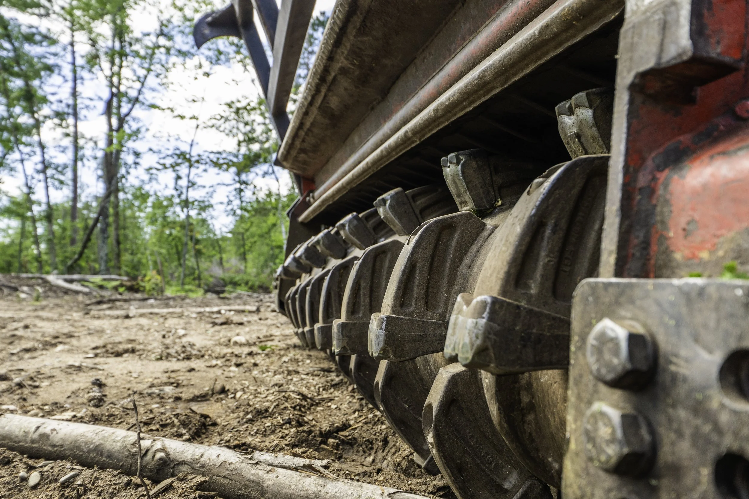 Close-up of a tracked vehicle's metal treads on dirt ground, with a background of green trees and blue sky.