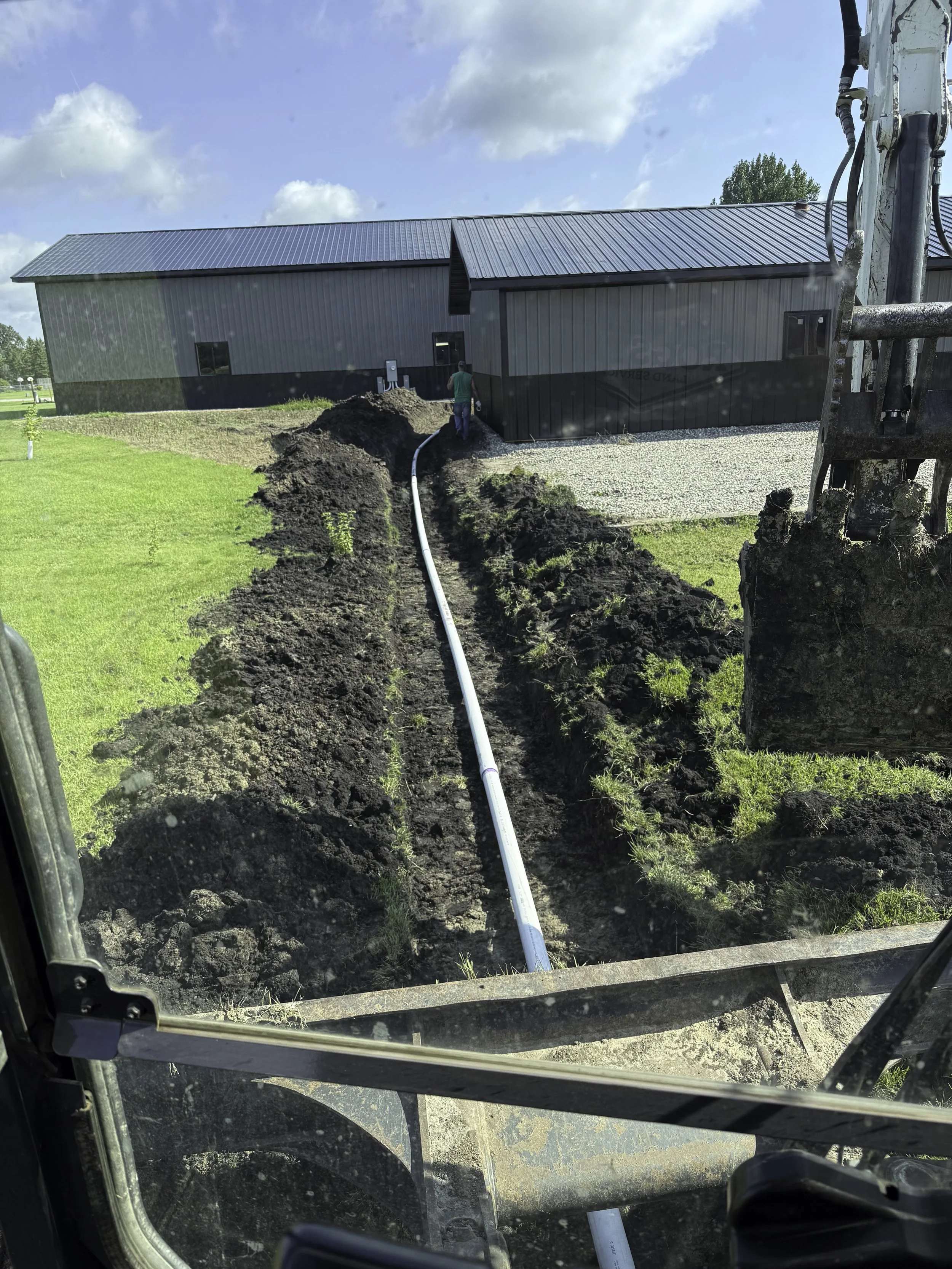 Construction worker operating machinery to install underground utility piping at a building site under a partly cloudy sky.
