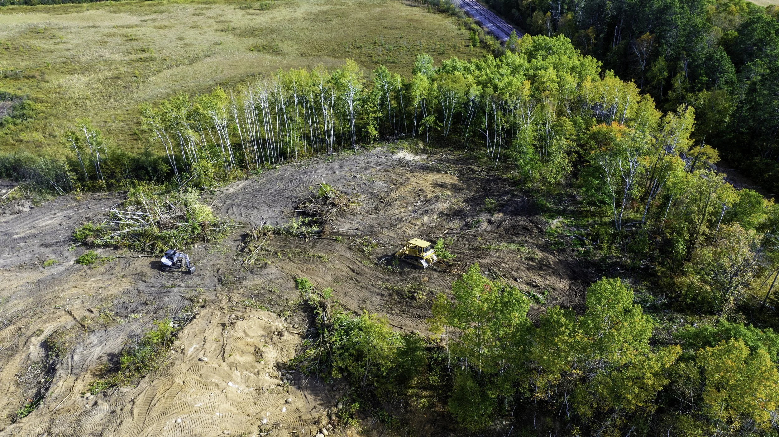 Aerial view of a cleared construction site with two heavy machinery vehicles surrounded by trees and a forest.