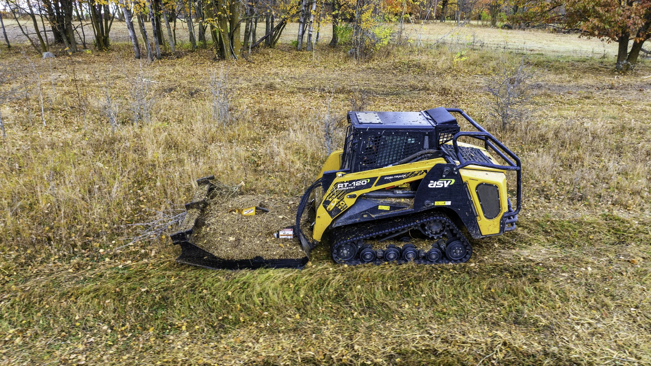 A small yellow and black tracked bulldozer pushing up dirt and small branches in a grassy, tree-filled area during autumn.