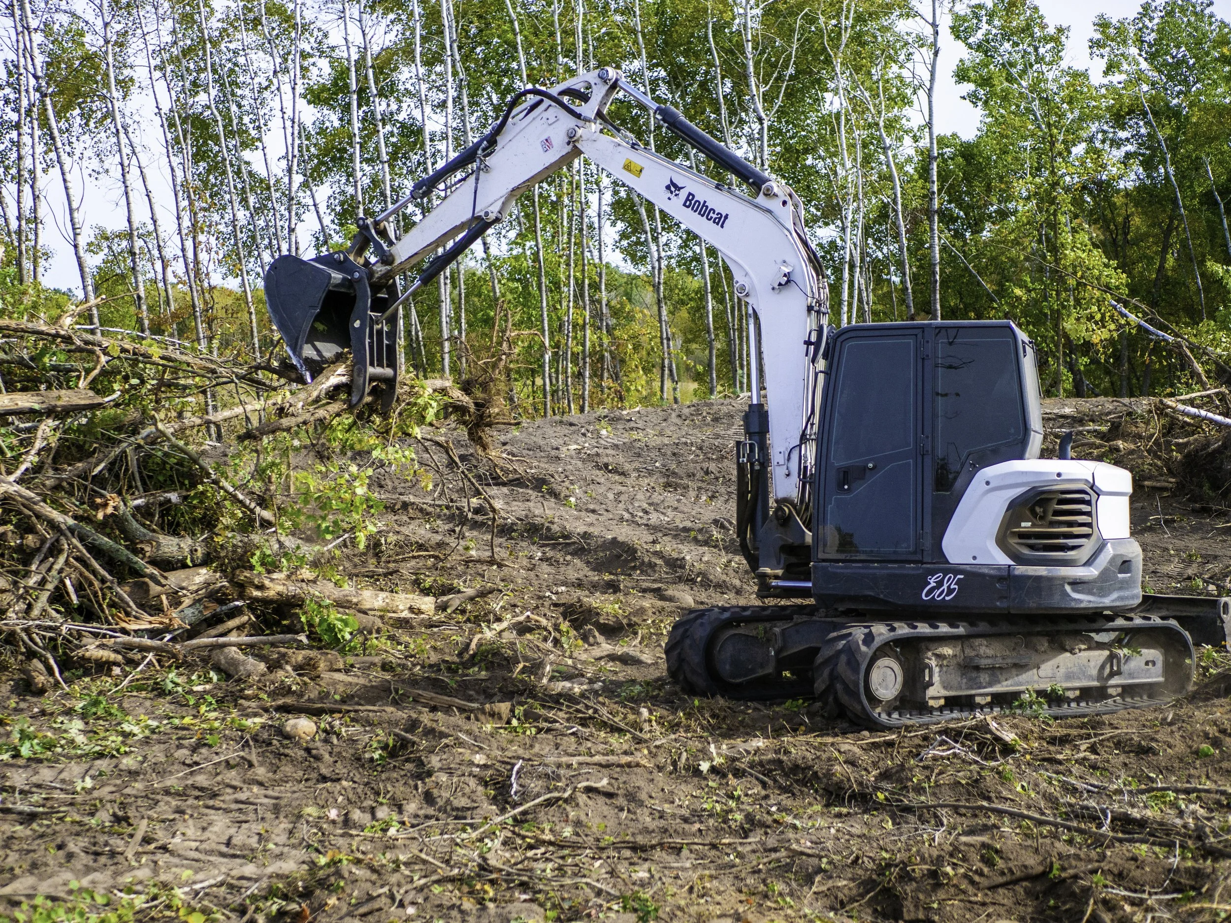 A small white and black excavator with the brand name Bobcat is clearing land by removing trees and tree debris in a forested area.