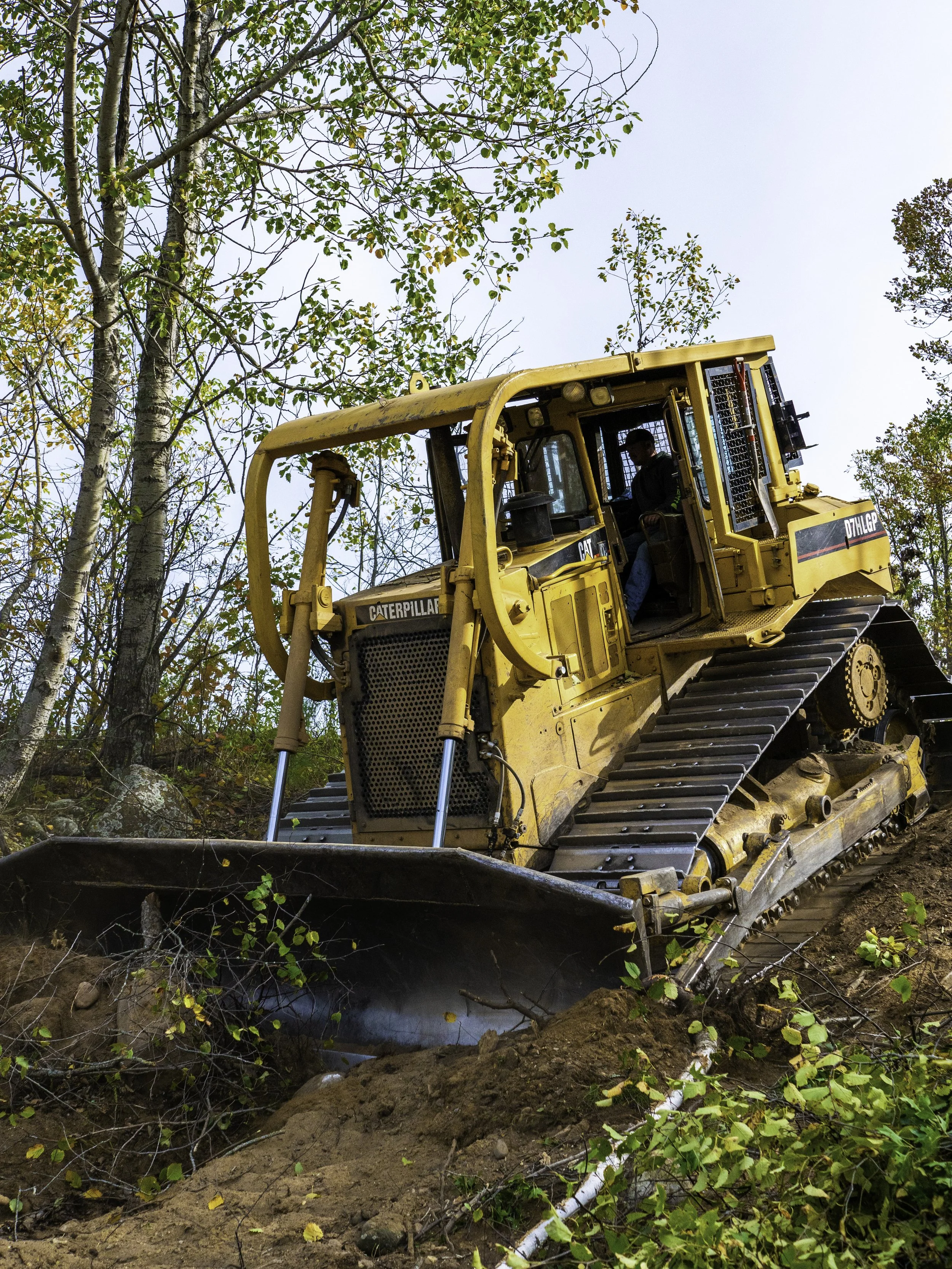 A yellow Caterpillar bulldozer working on a dirt slope, surrounded by trees with changing leaves, under a clear sky.