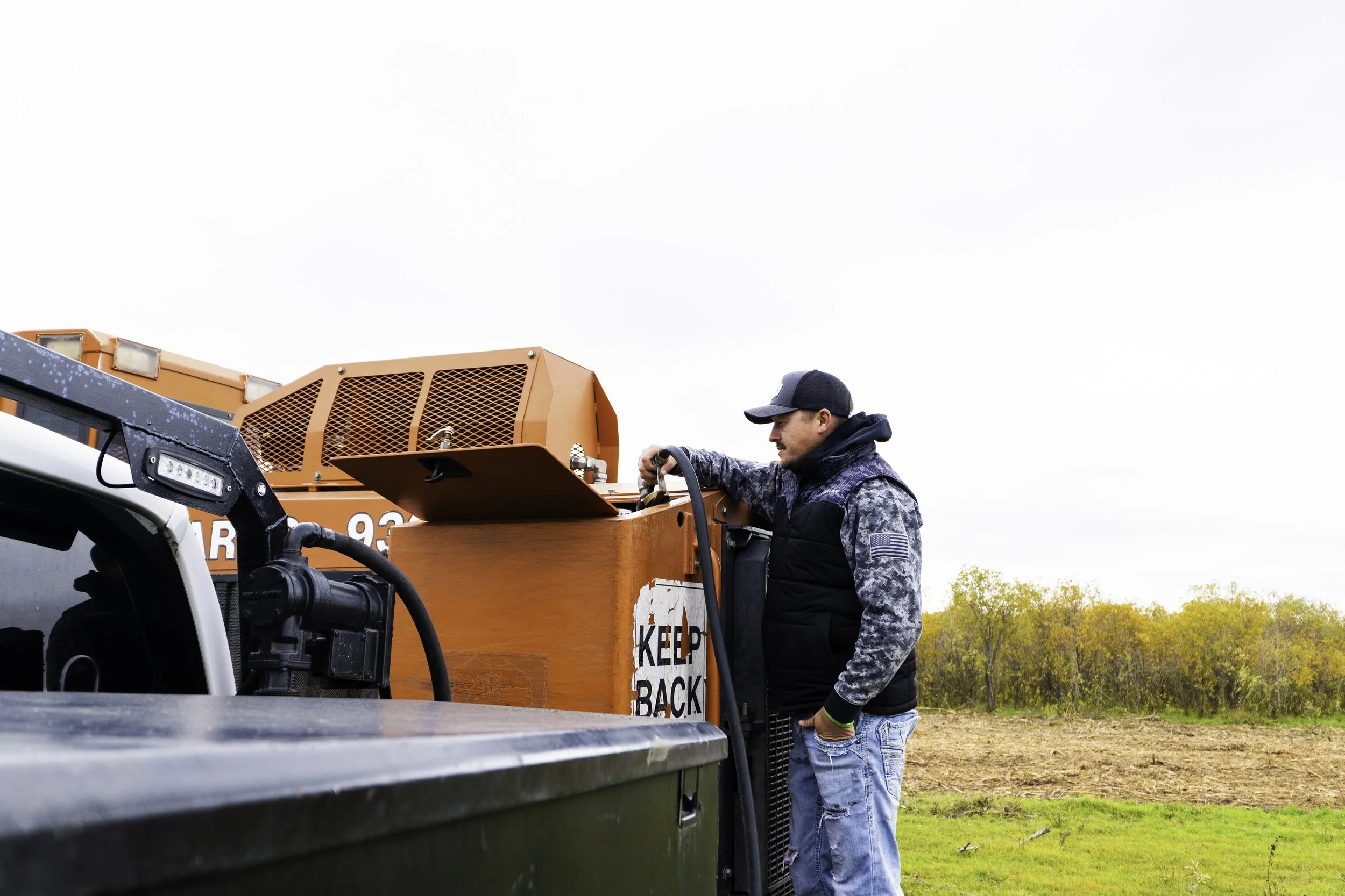 A man in camouflage and black jacket filling a large orange generator with fuel in an open field.