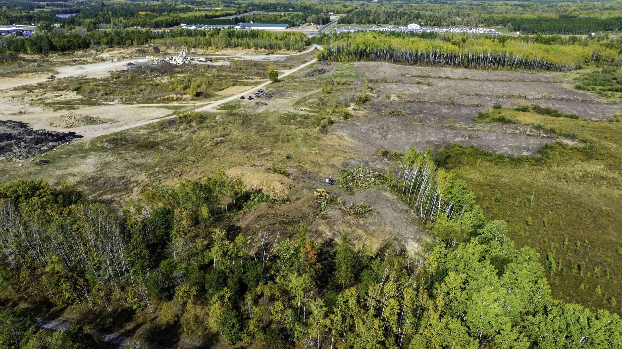 An aerial view of a construction or land clearing site with trees, dirt roads, and heavy equipment, with a forested area in the background.