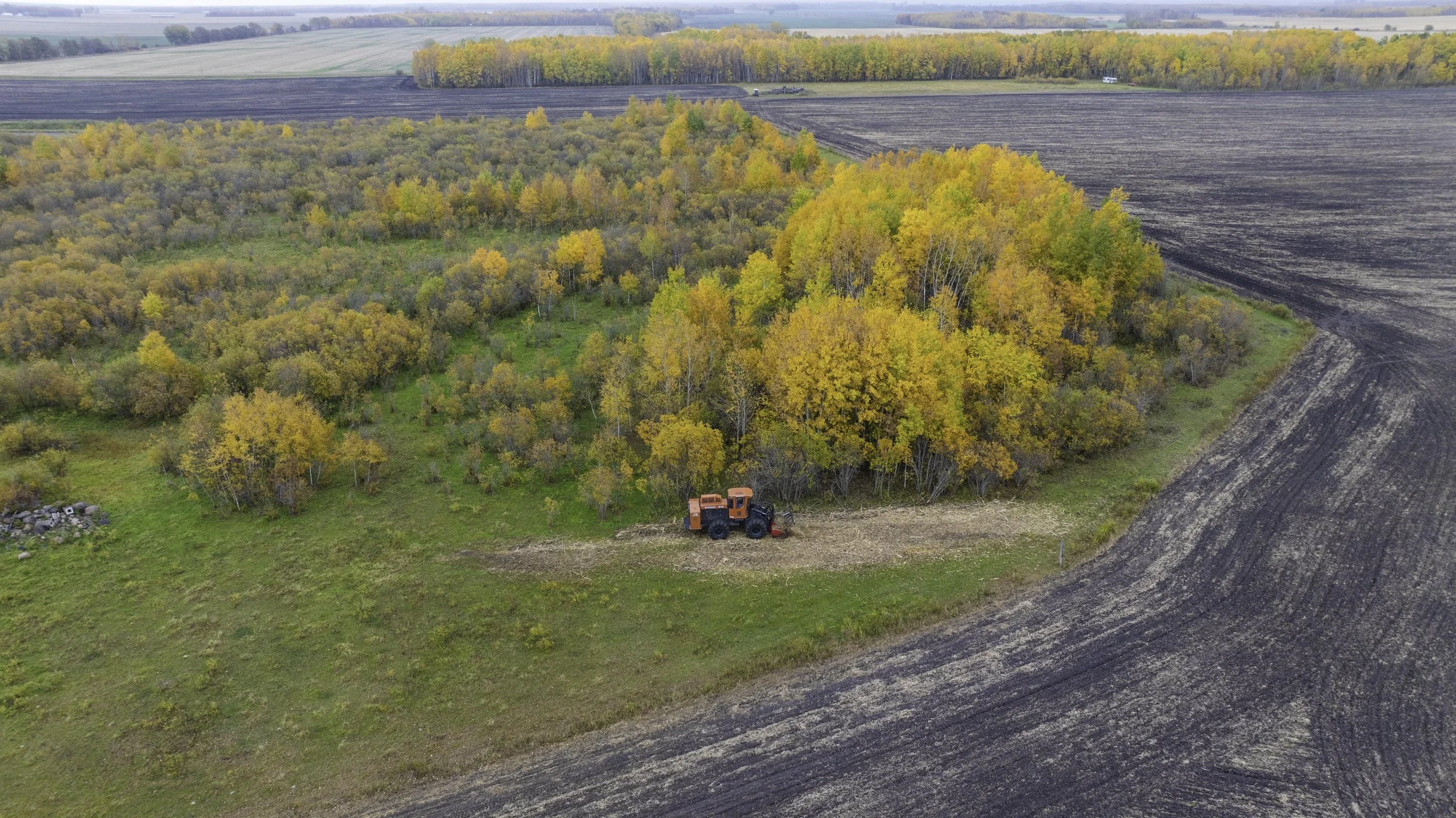 An aerial view of a farm field with a tractor and fall-colored trees. The tractor is near the edge of a small tree grove, with harvested land and distant fields in the background.
