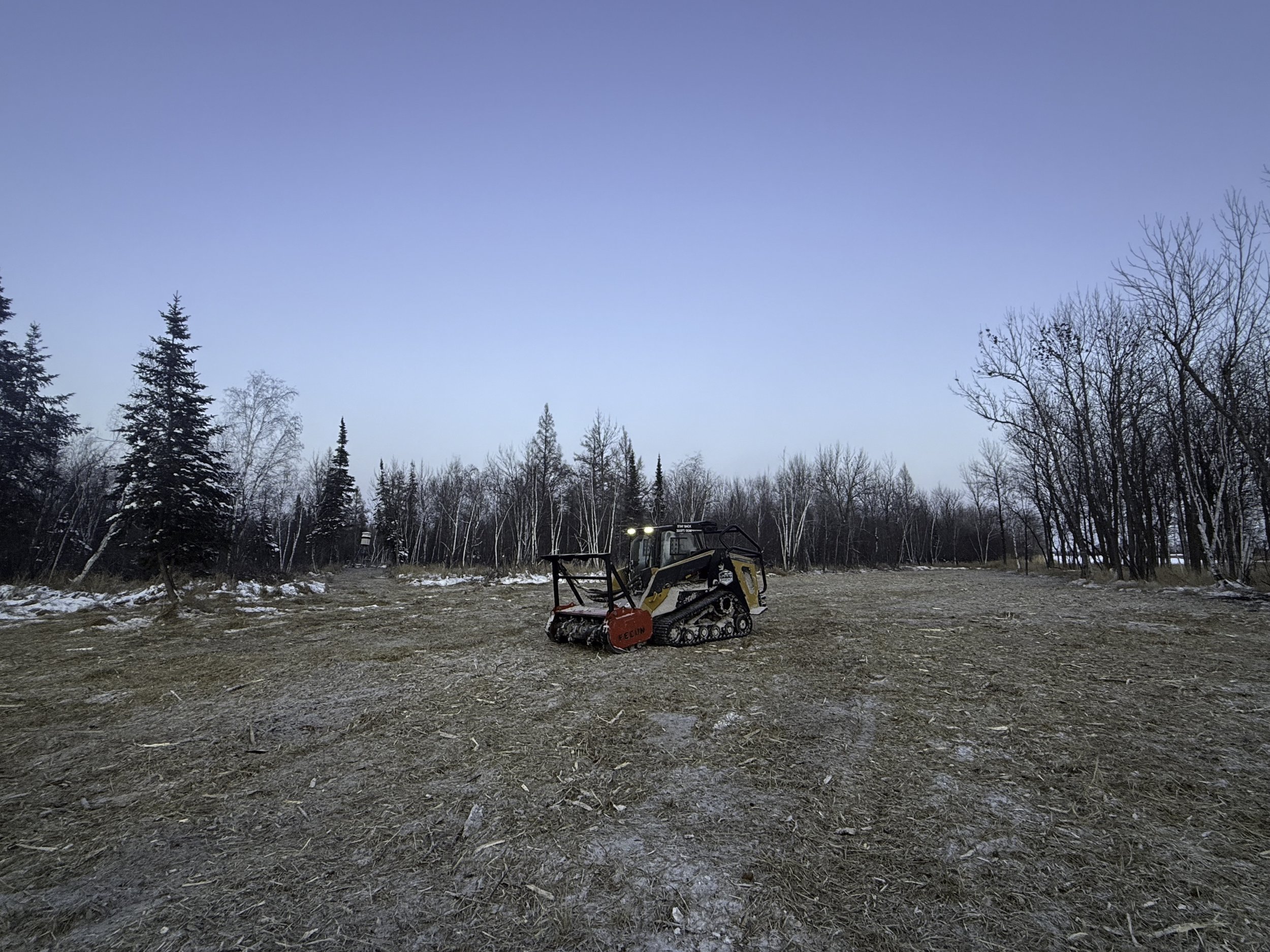 A tractor on a mostly cleared open field with some snow, surrounded by leafless and evergreen trees under a clear blue sky.