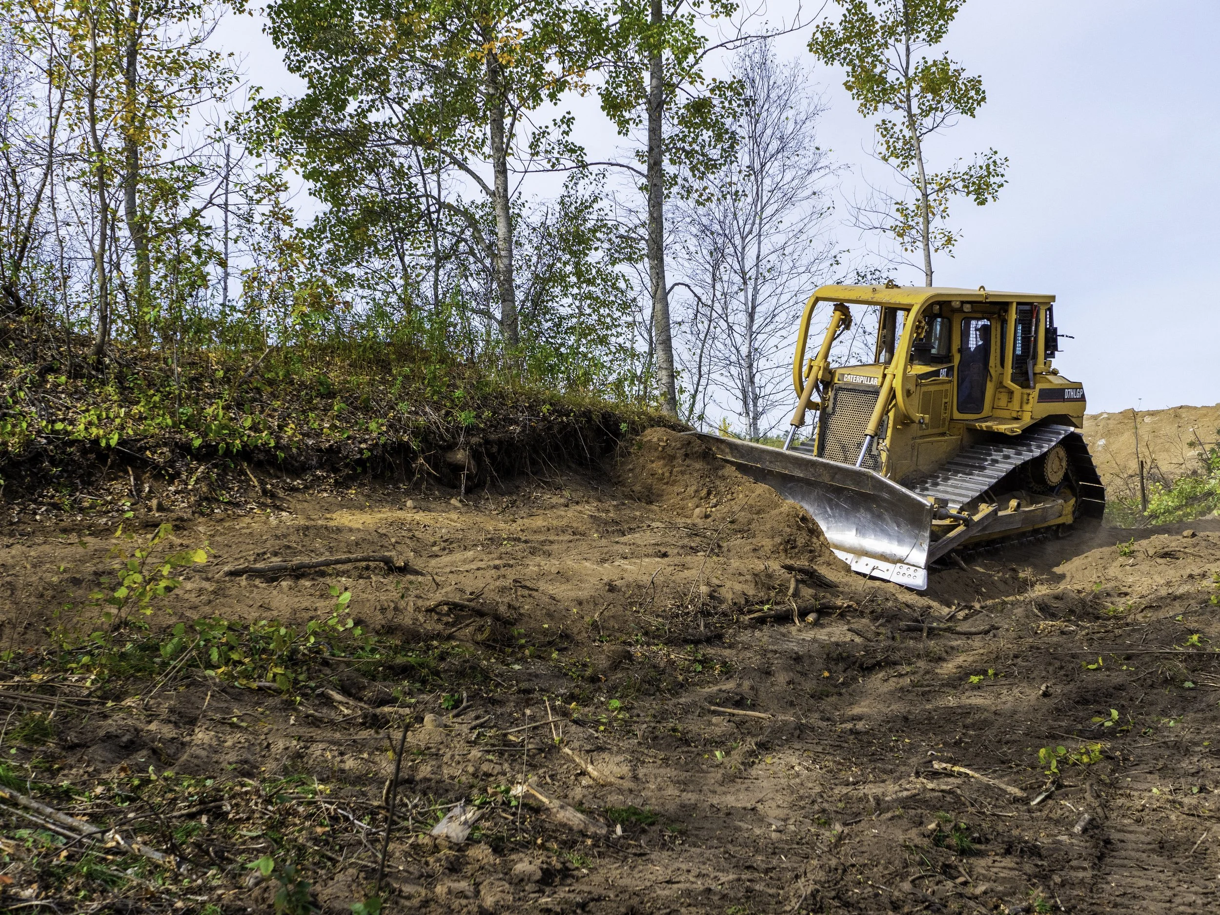 A yellow bulldozer working on a dirt hill with trees in the background.