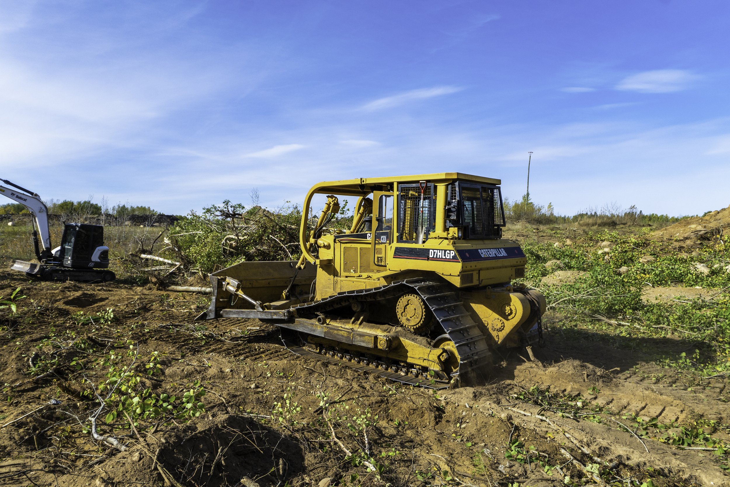 A yellow Caterpillar bulldozer operating on a dirt field with some green bushes and a clear blue sky in the background.