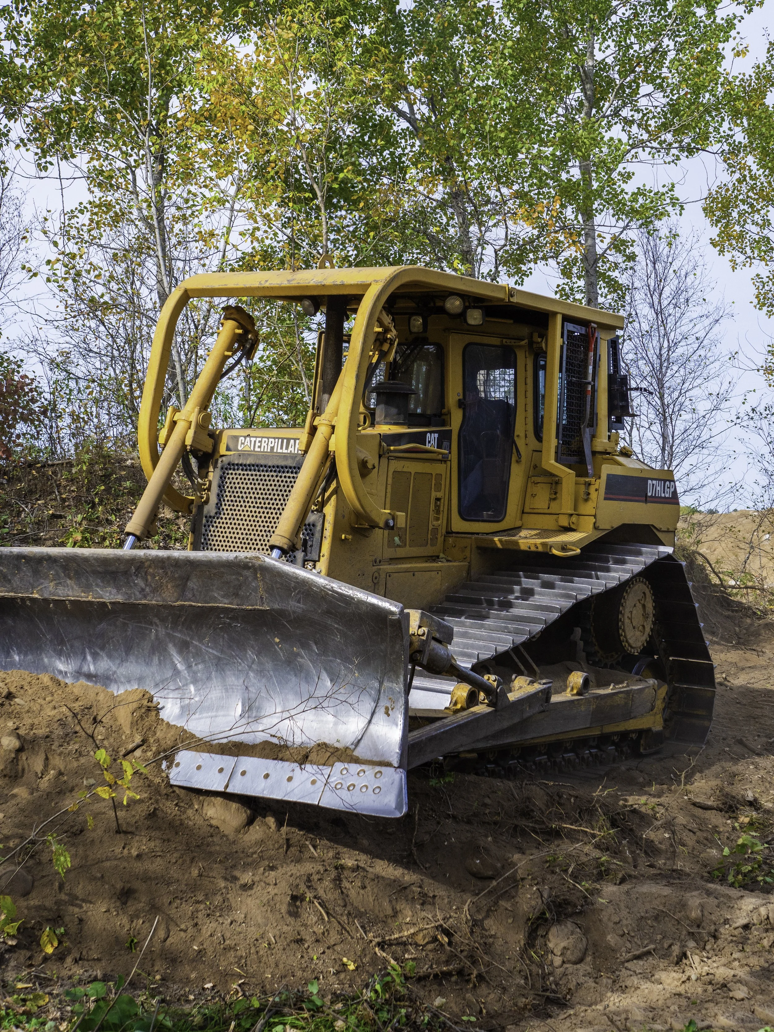 A yellow Caterpillar bulldozer on a construction site surrounded by dirt and trees with green and some yellow leaves.