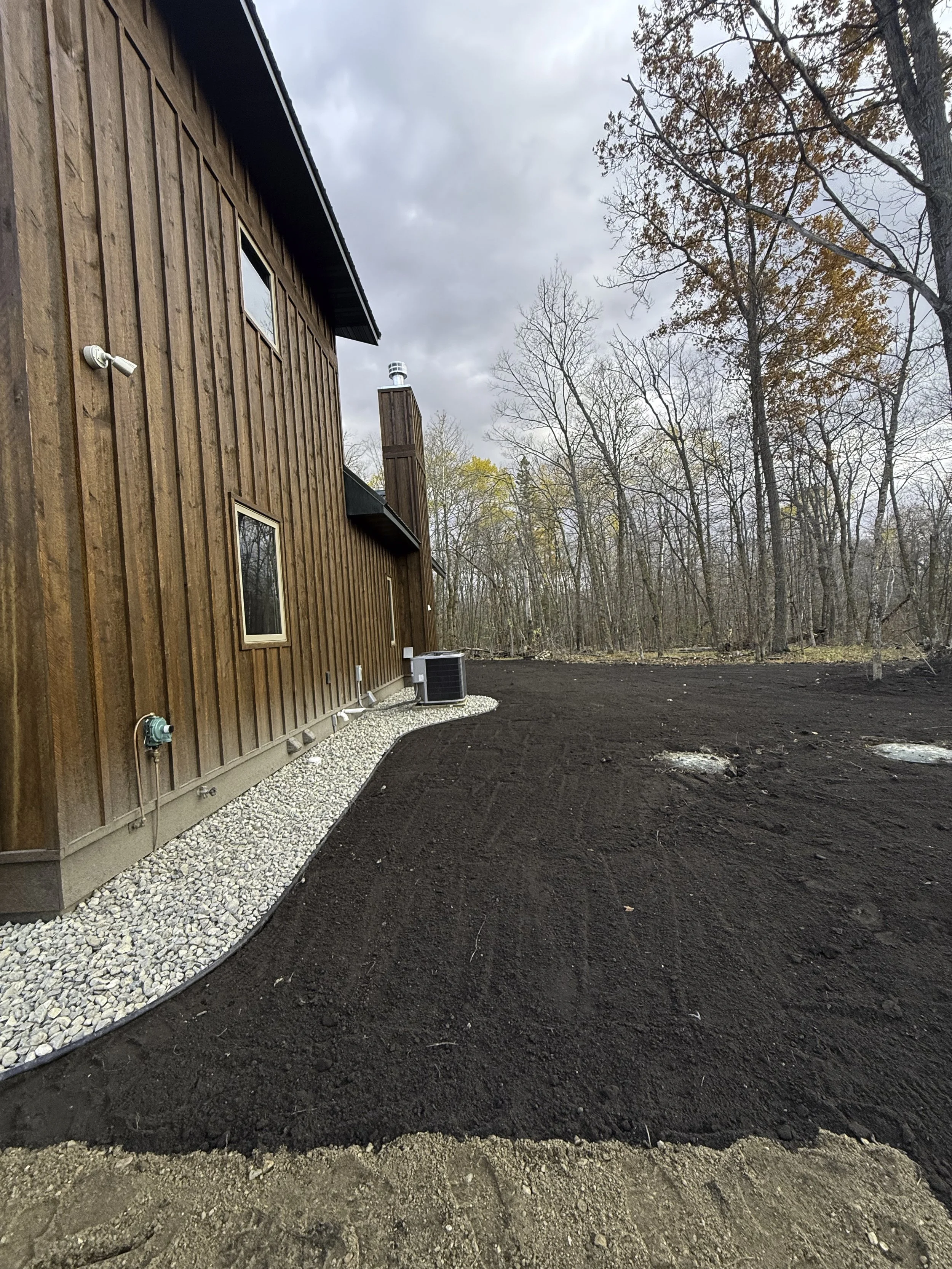 Side view of a house with a wooden exterior, a small window, and a chimney, surrounded by freshly tilled dark soil and a gravel pathway, with leafless trees in the background and cloudy sky overhead.