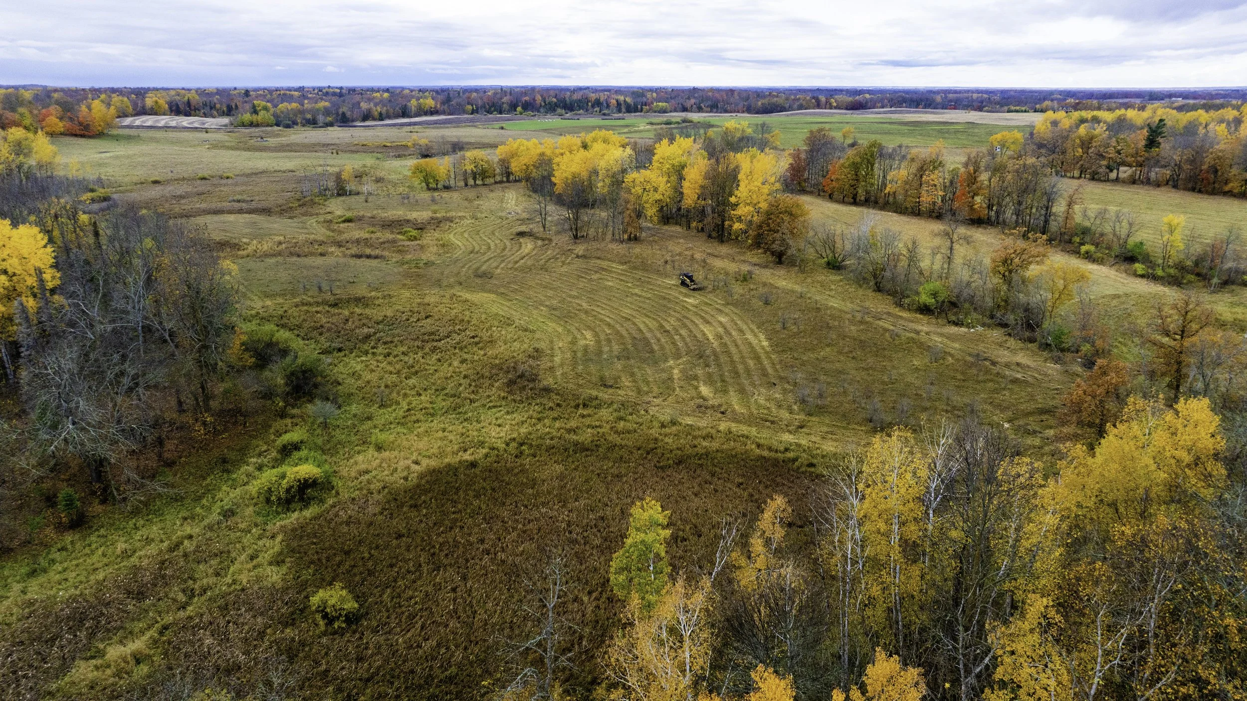 Aerial view of a countryside landscape in autumn with colorful trees, open fields, and a tractor.