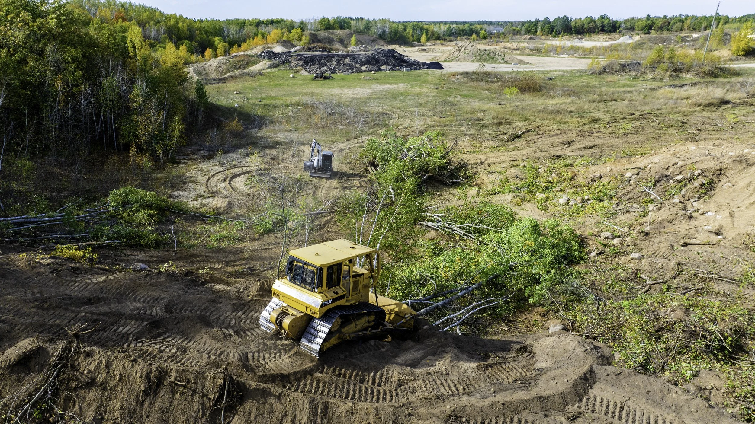 Construction equipment clearing fallen trees and debris from a large, open dirt area in a forested landscape.