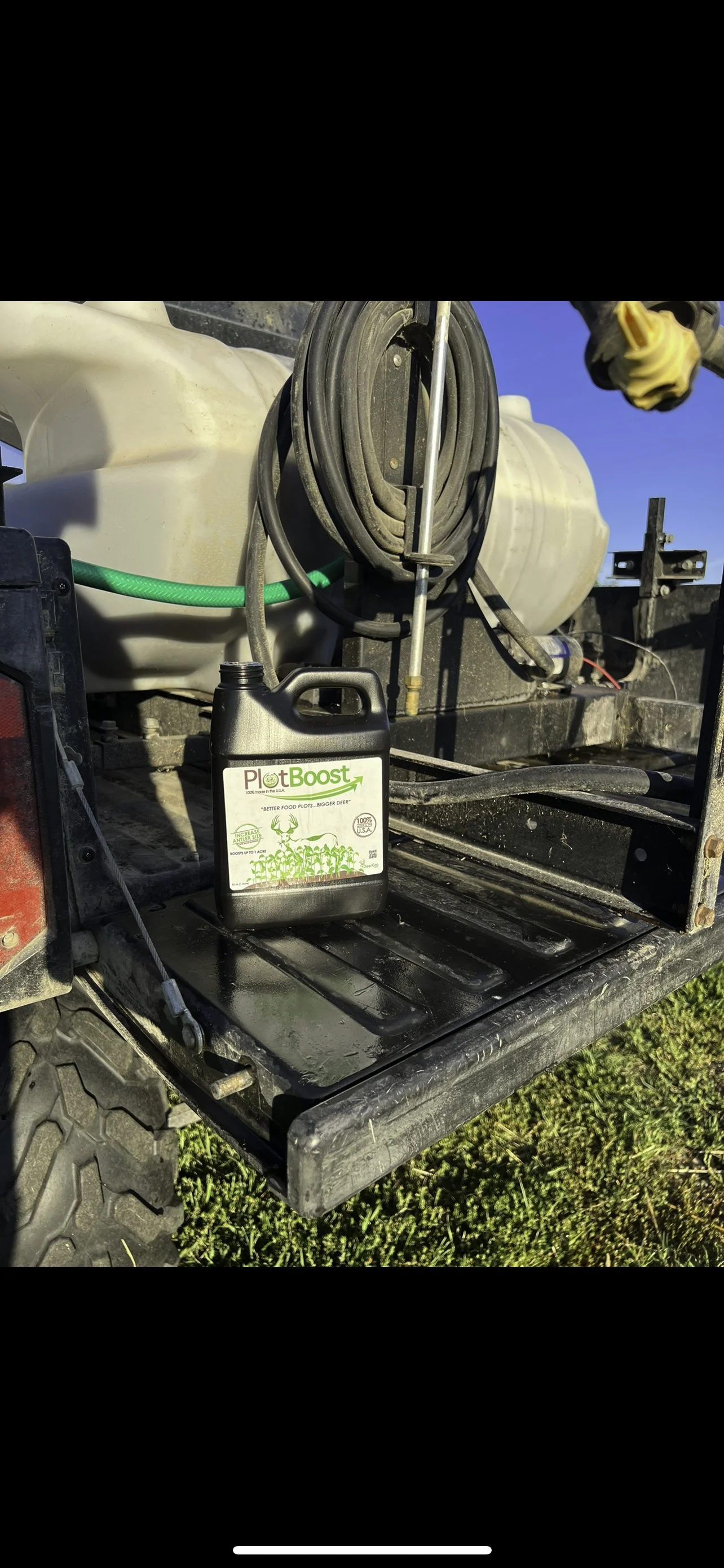 A black container labeled Plant Boost with a green and white label sitting on a metal surface, with hoses and a large white tank in the background.
