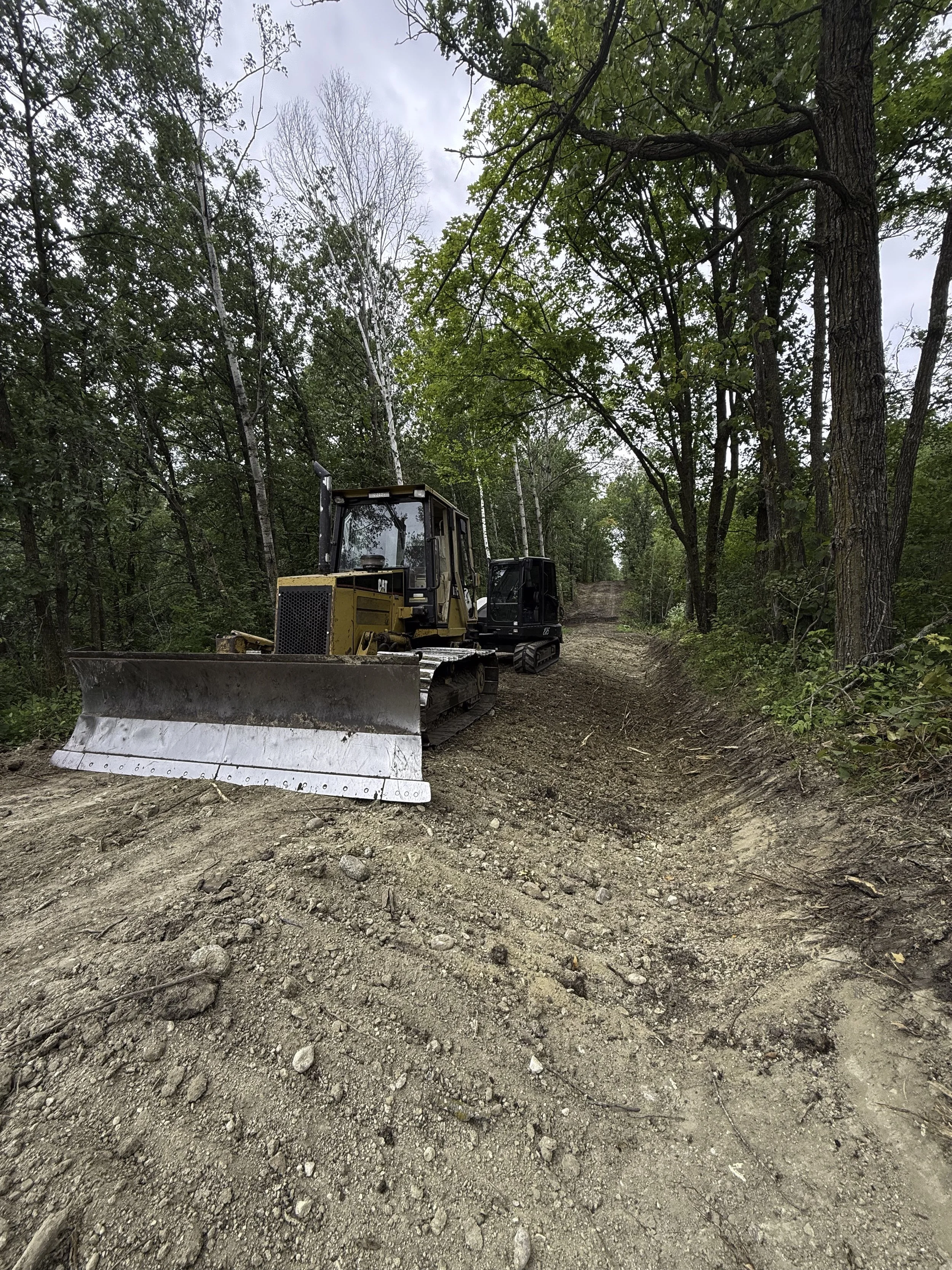 Construction machinery on a dirt trail in a forest, with trees on both sides and a cloudy sky overhead.