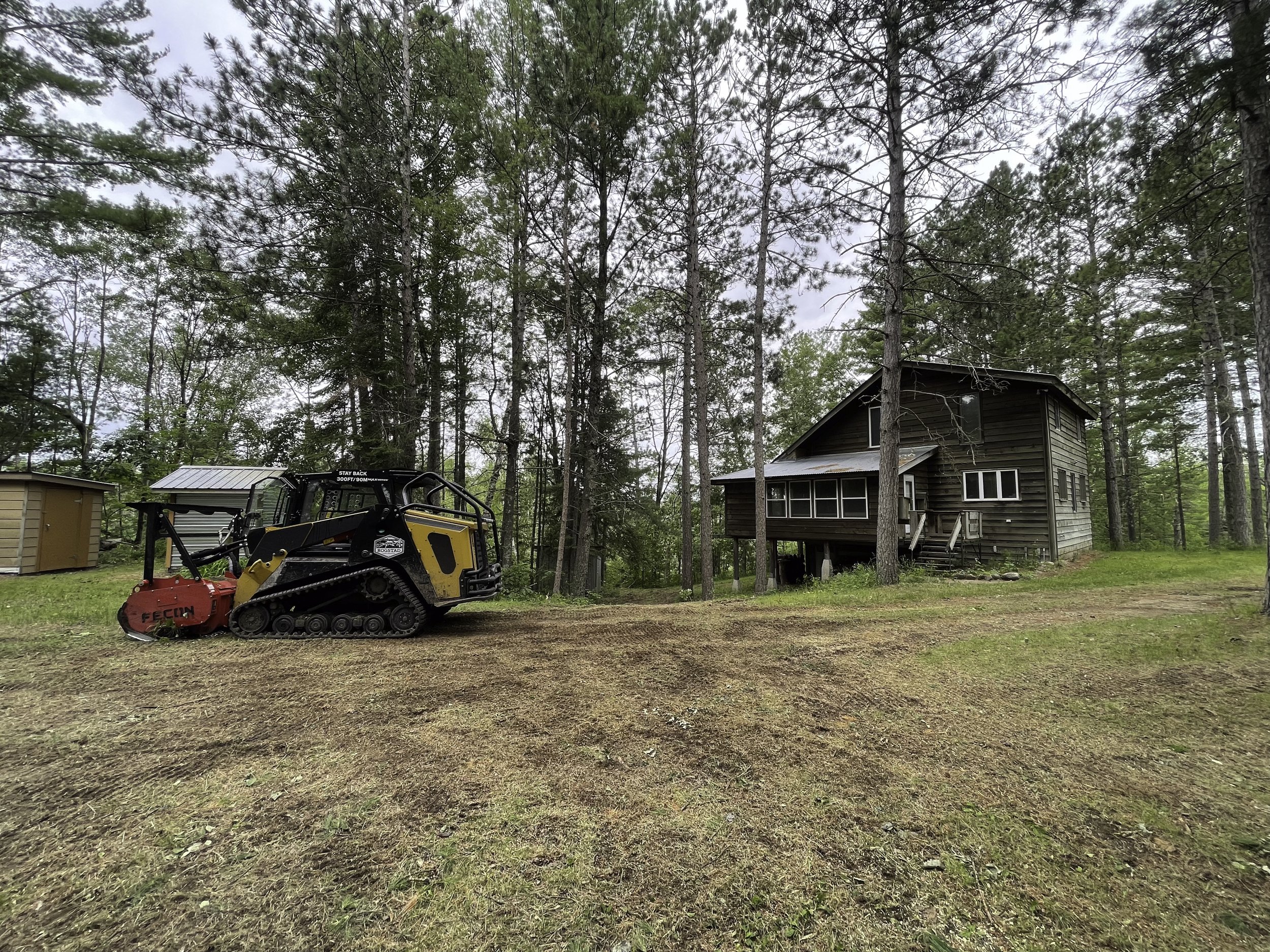 A wooded yard with a small brown house on stilts, a red and black tracked compact utility loader, and a small brown shed in the background.
