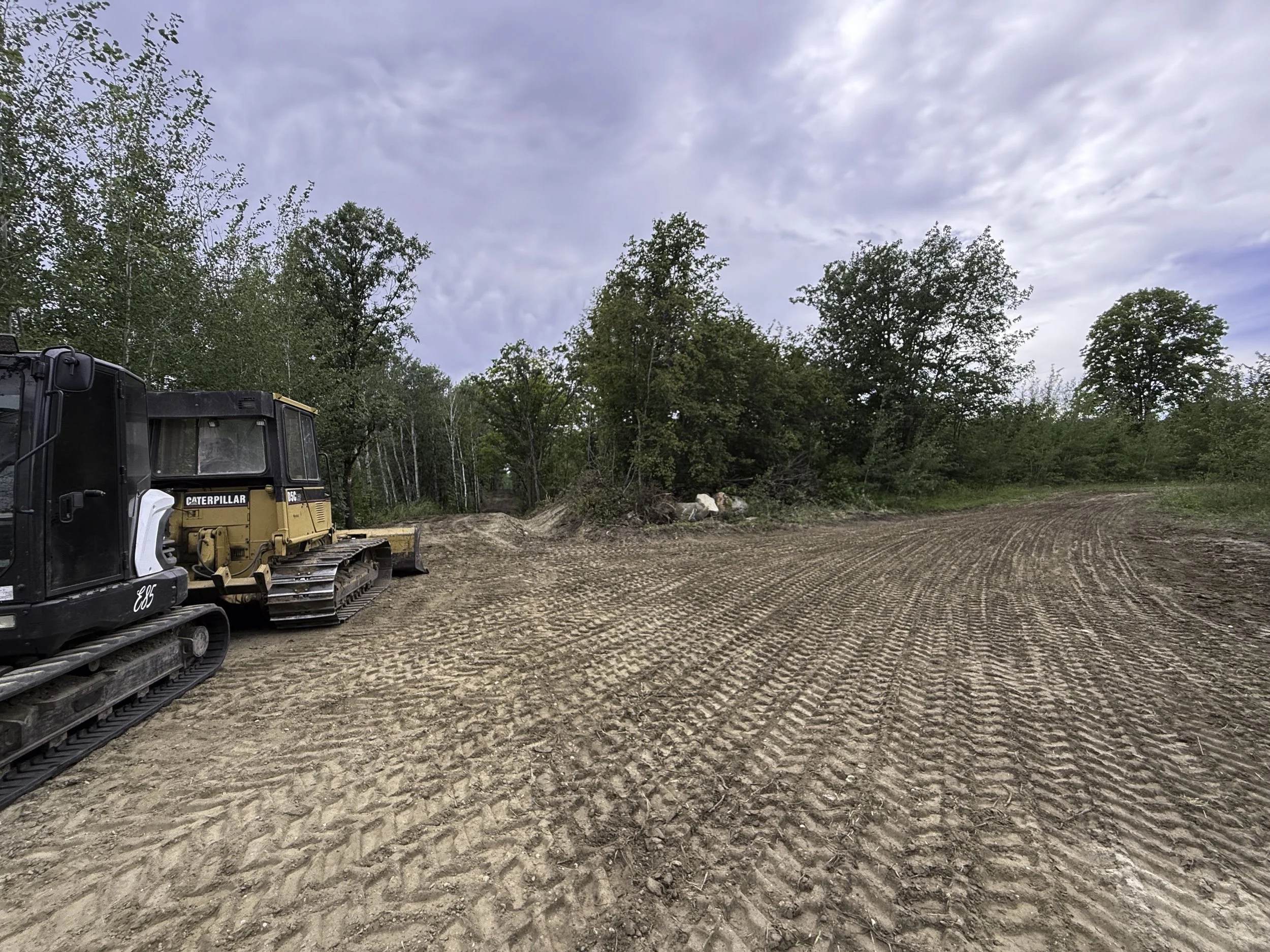 A construction site with a Caterpillar bulldozer parked on dirt with tire tracks, surrounded by trees under a cloudy sky.