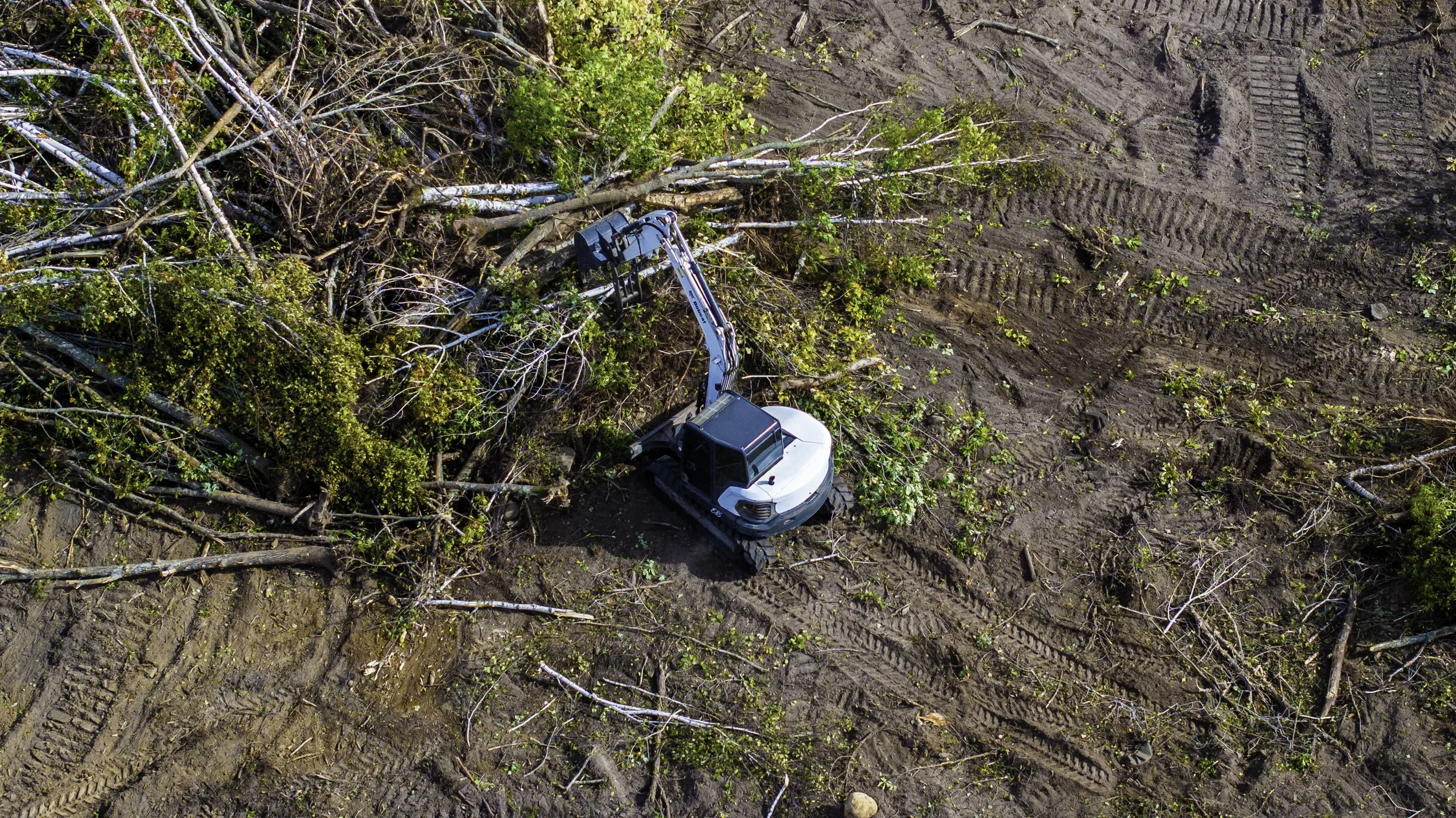 An aerial view of a construction site with a white excavator clearing fallen trees and branches among disturbed soil with tire tracks.