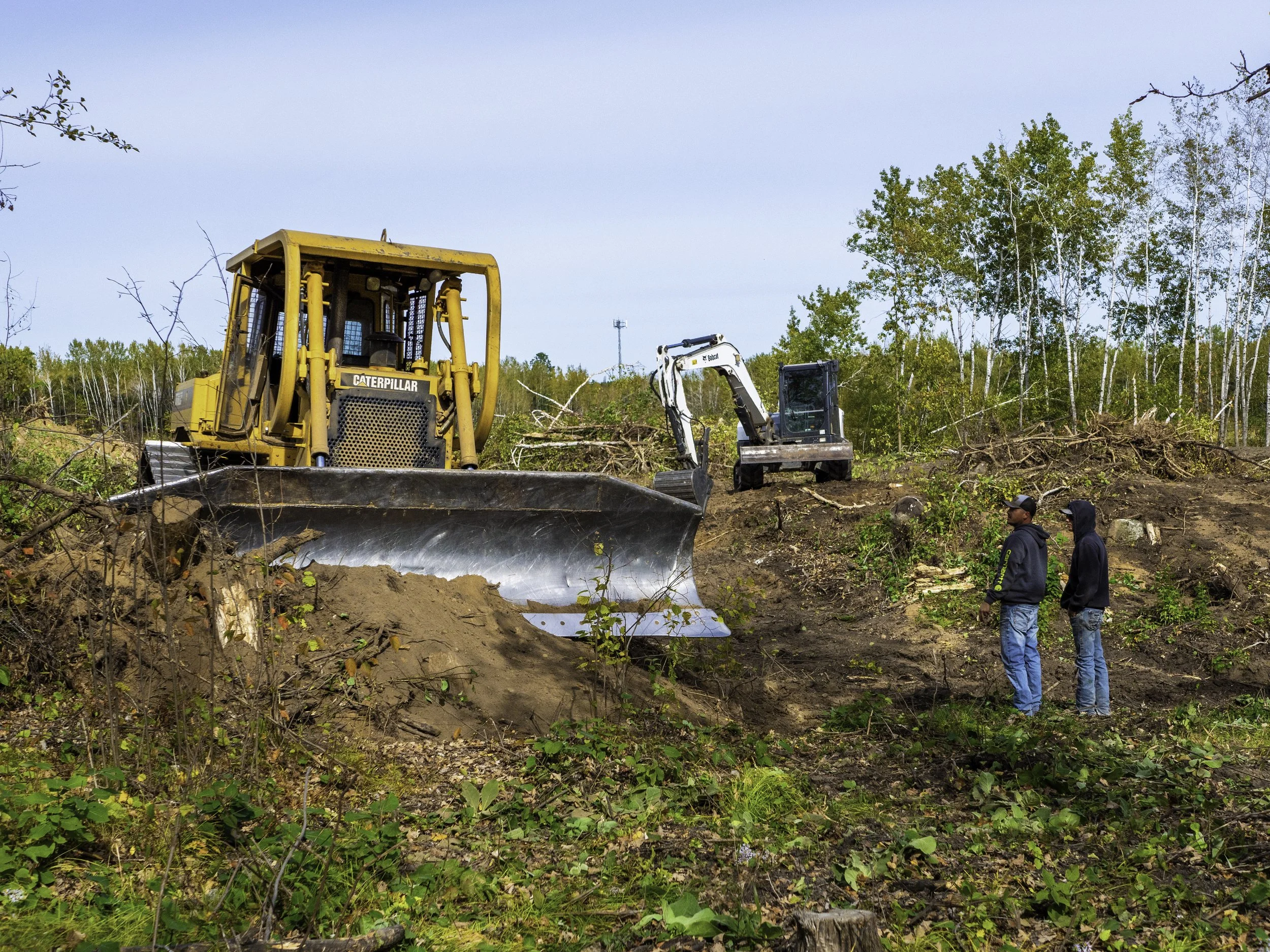 Two men stand on a cleared patch of land with dirt and small plants, observing construction equipment including a yellow bulldozer and a white excavator, with trees in the background under a clear sky.