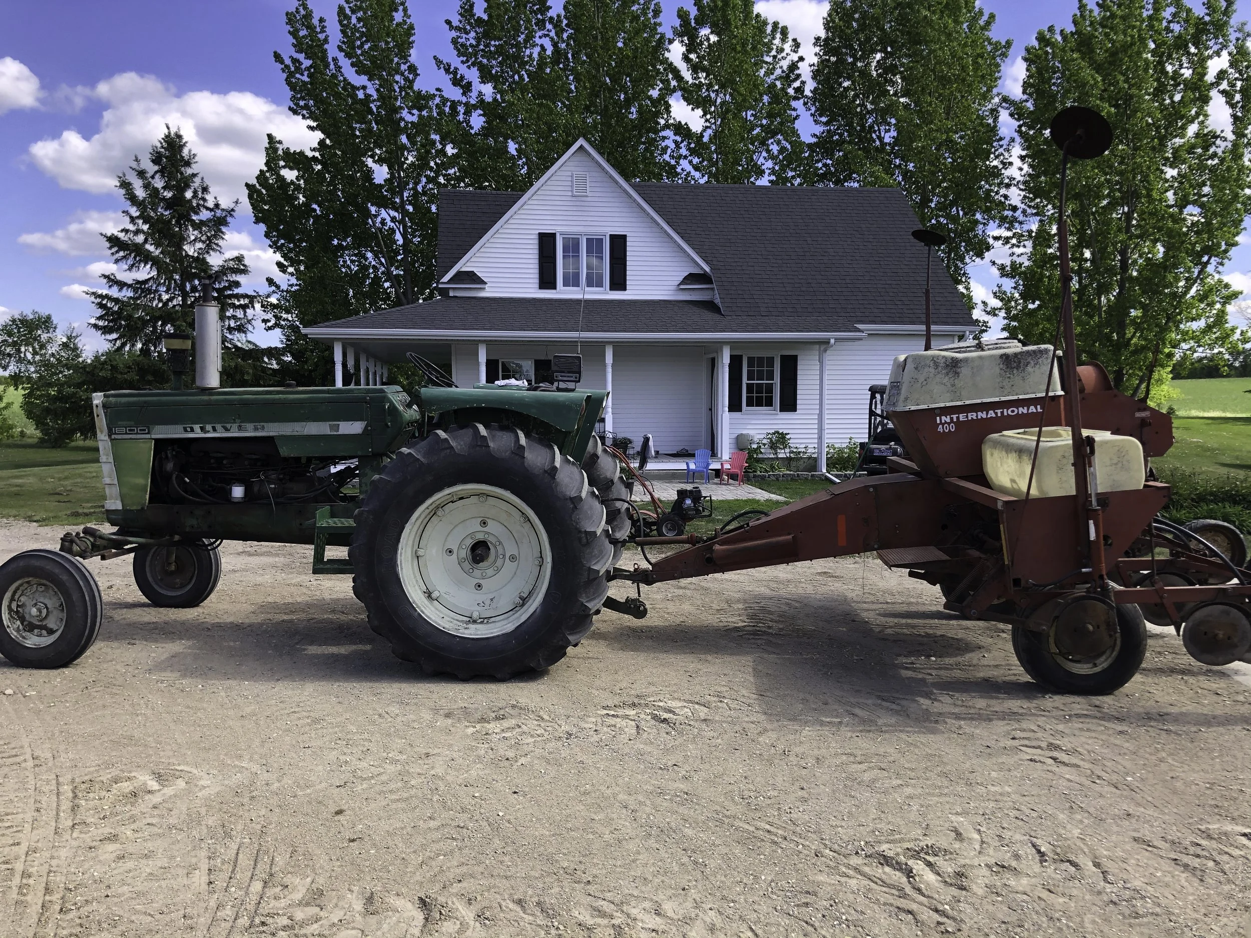 A green Oliver tractor attached to a red International 400 hay rake in a rural yard with a white house and green trees in the background.