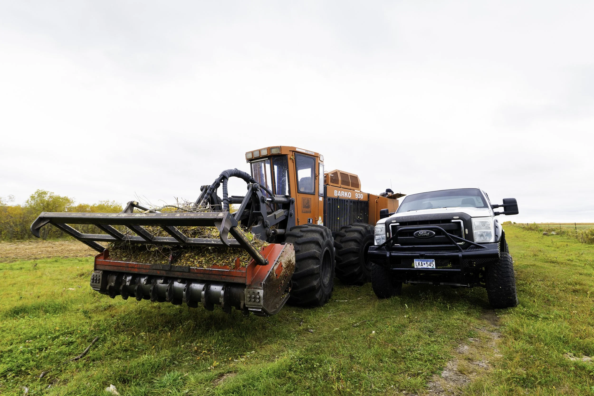 An orange BARKO tractor with large black tires and a front attachment for clearing debris, parked next to a black Ford truck with large tires on a grassy field with yellow and green plants in the background and overcast sky.