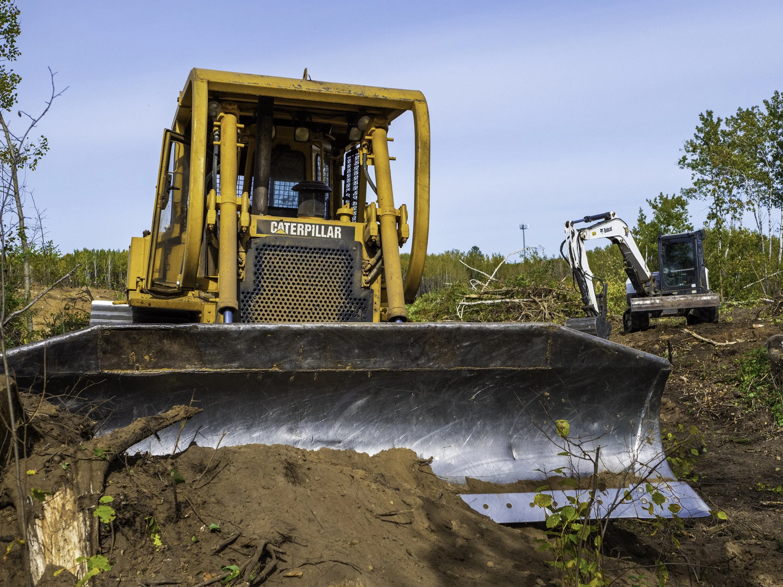 A large yellow Caterpillar bulldozer and a smaller white micro excavator working on a dirt landscape with trees and a partly cloudy sky in the background.