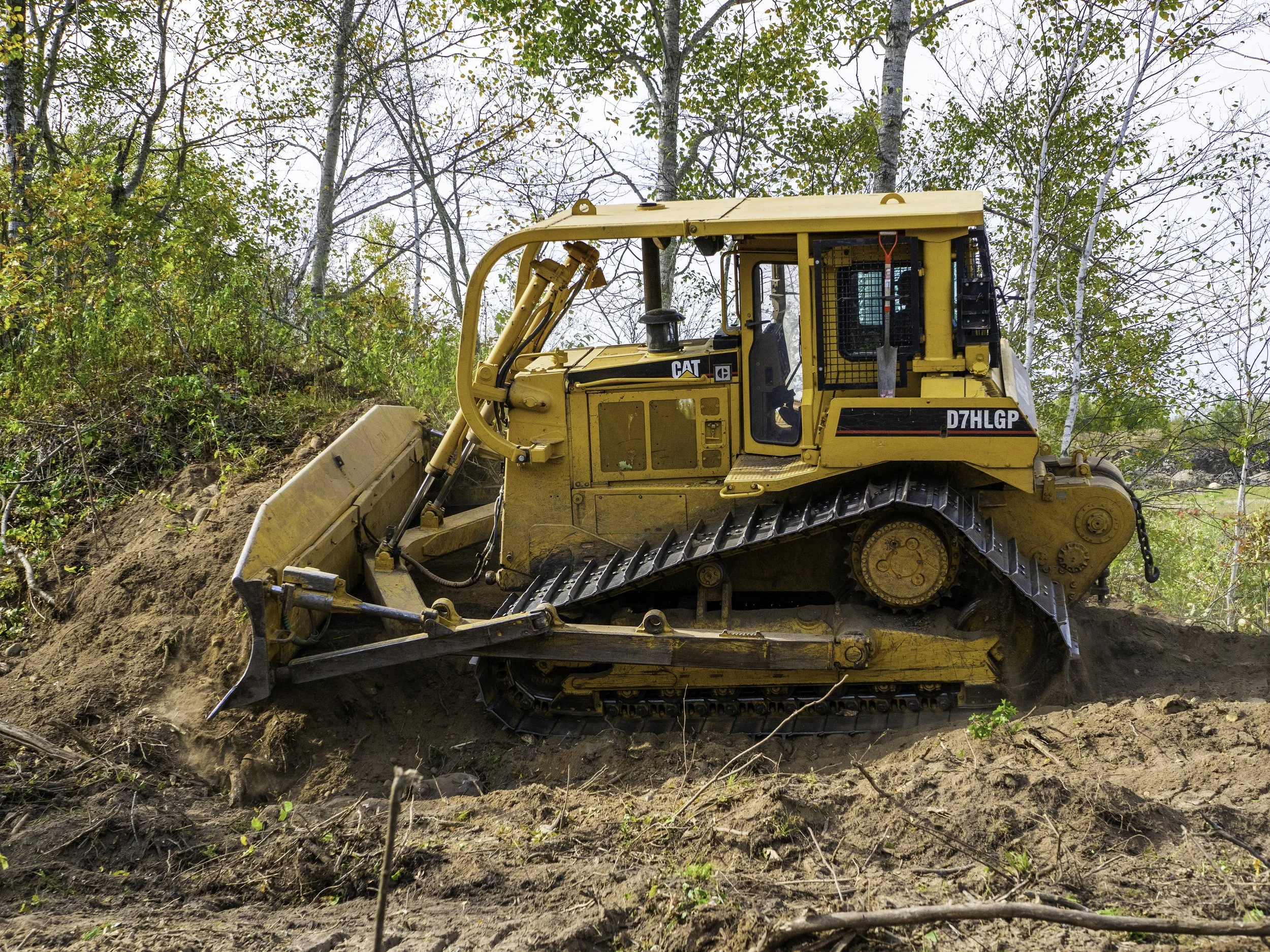 A yellow bulldozer working on a dirt hill in a wooded area with trees and foliage in the background.