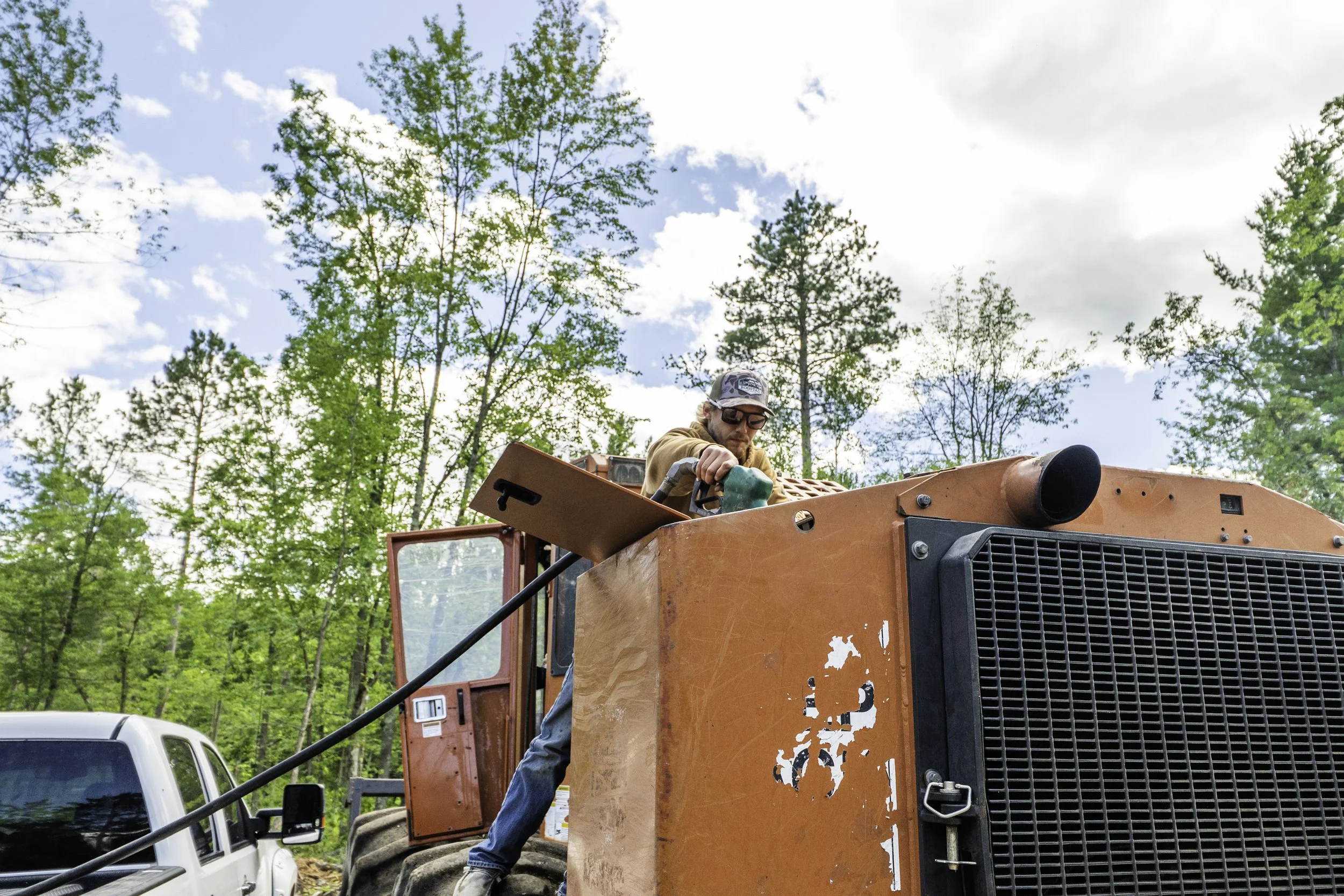 A man wearing sunglasses, a baseball cap, and a brown jacket is working on an orange construction vehicle in a forested area with green trees and a partly cloudy sky.