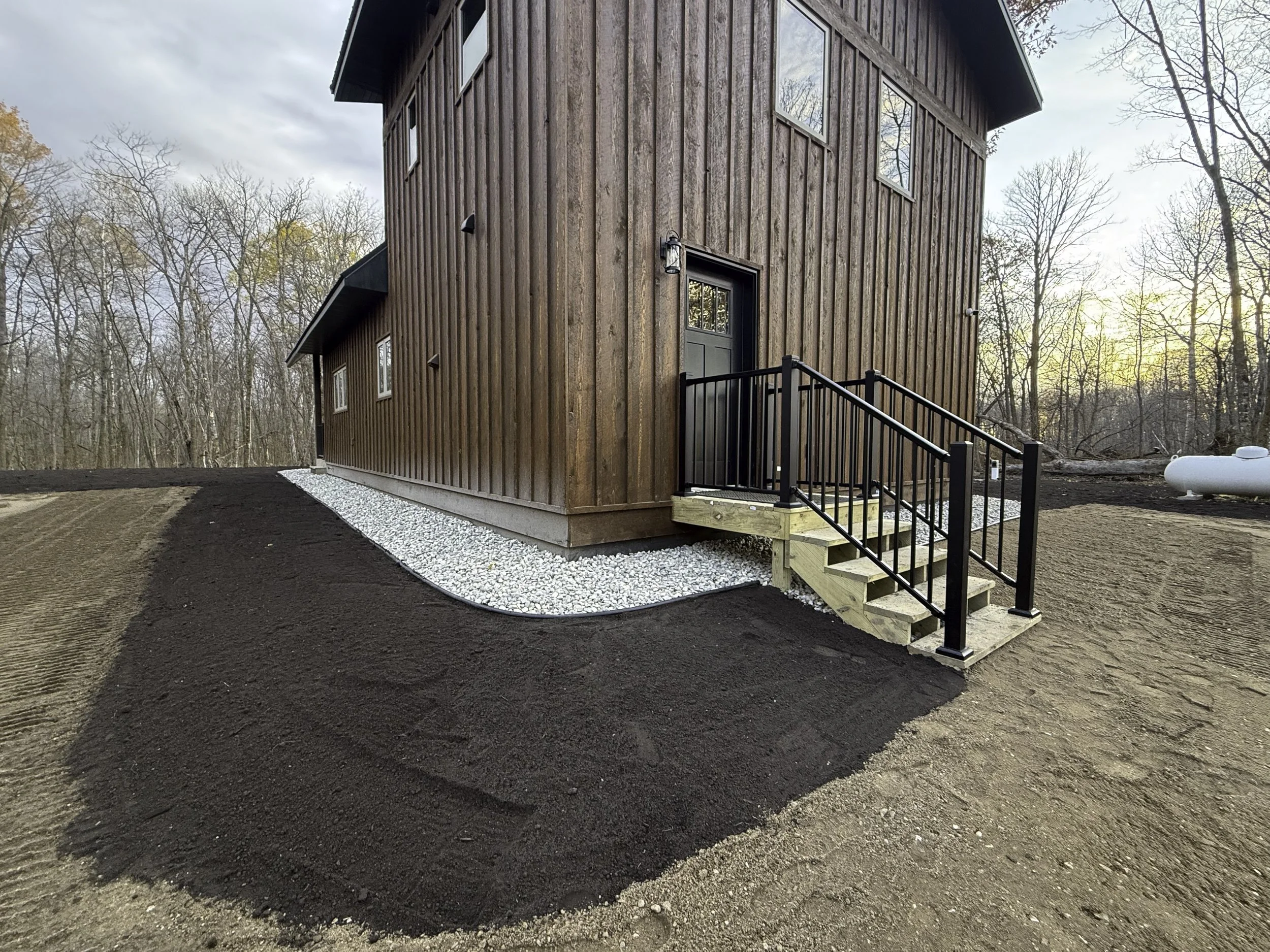Newly constructed wooden house with a small staircase and black railing, surrounding dirt and gravel landscaping, in a wooded area at sunset.
