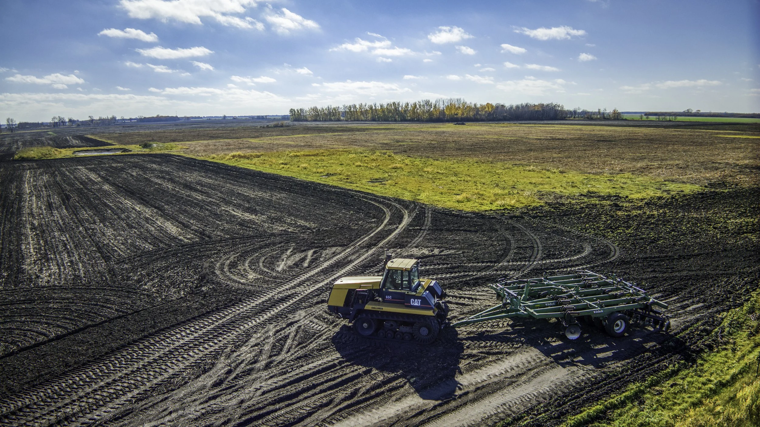 Aerial view of a yellow and blue CAT tractor working on black soil in a large open farmland under a partly cloudy sky.