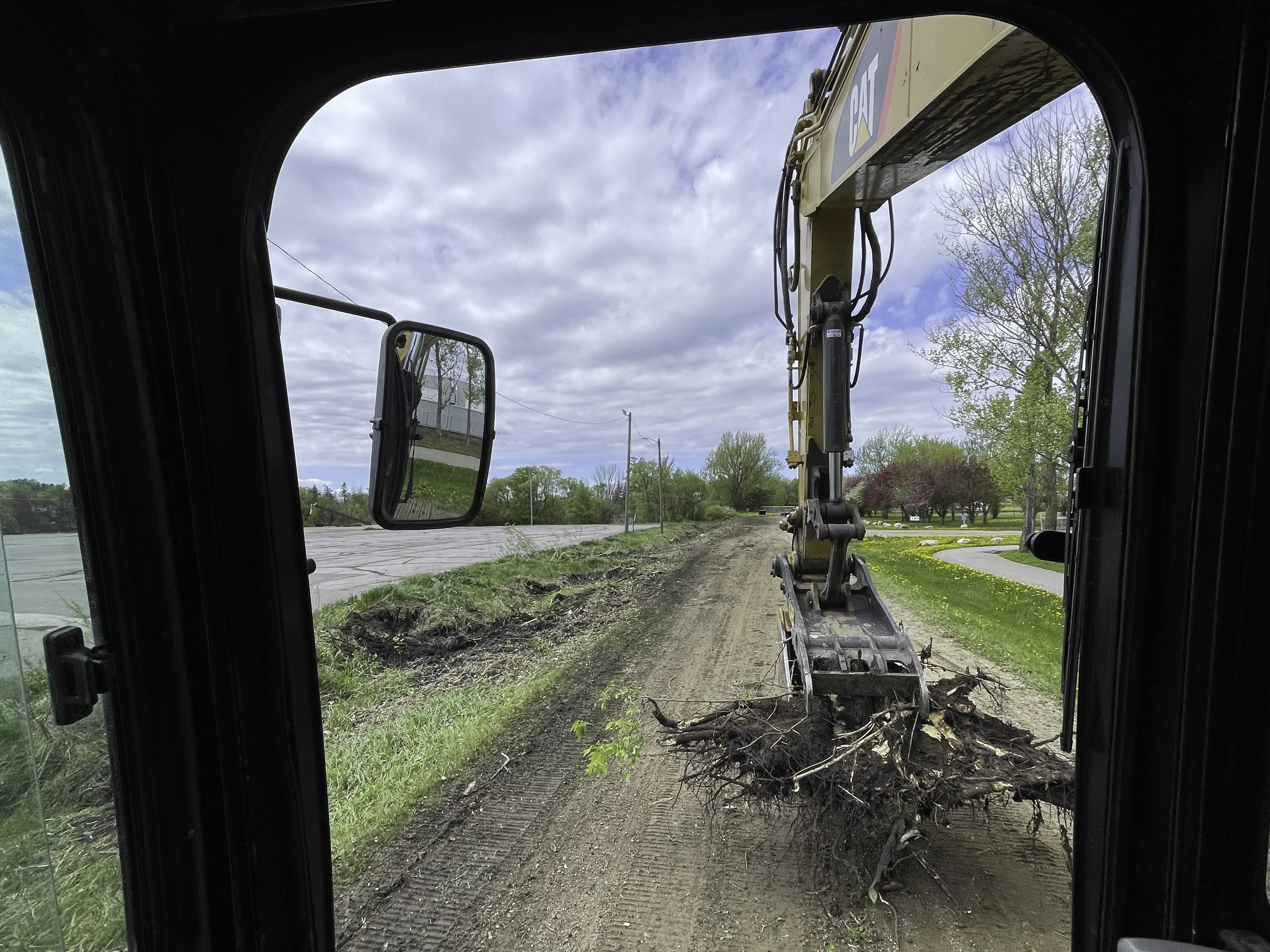 View from inside a construction vehicle, looking out at a dirt path being cleared with a backhoe. There are trees, grass, and a cloudy sky in the background.