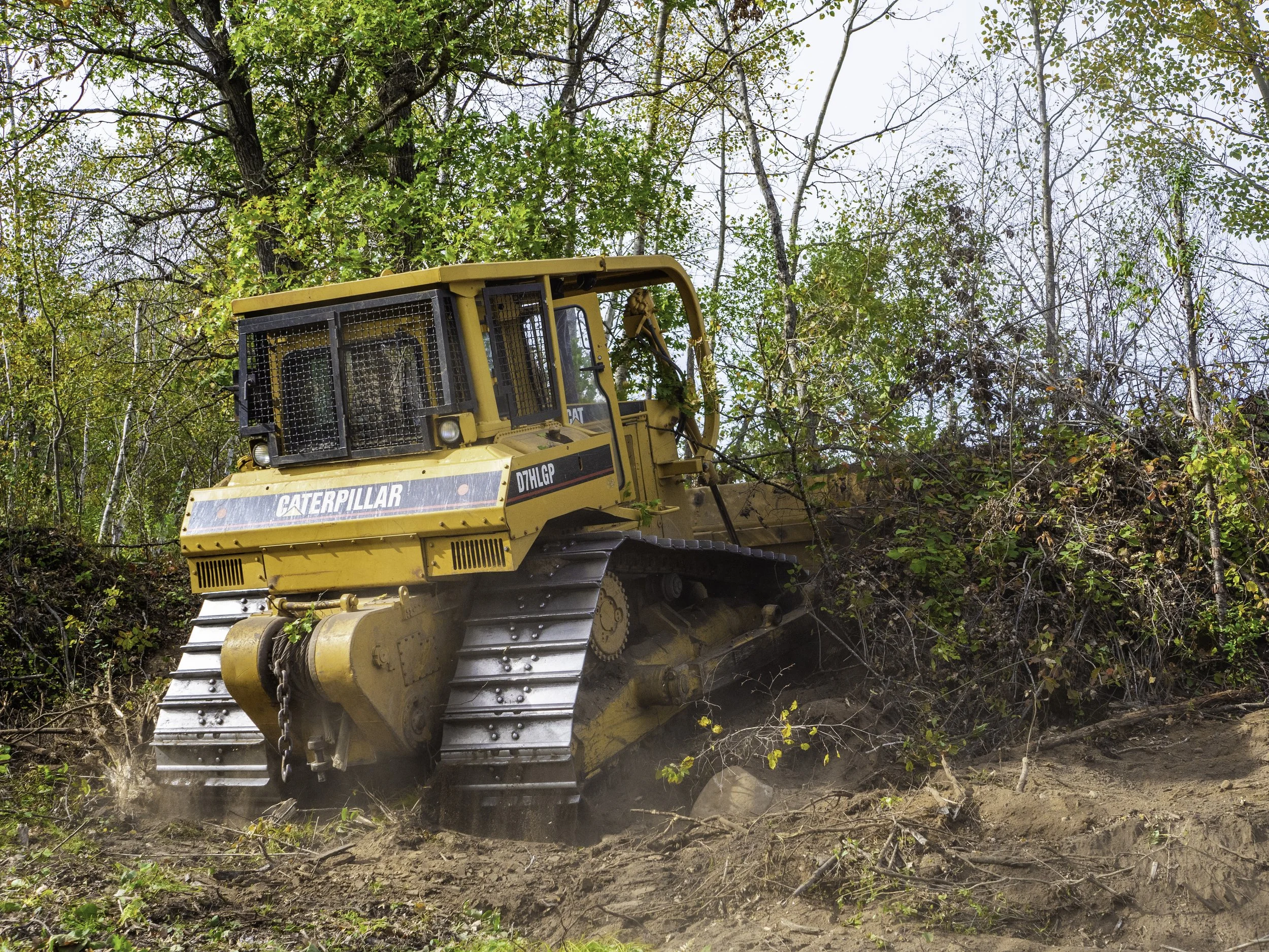 A yellow Caterpillar bulldozer pushing dirt and clearing brush in a wooded area with trees and green foliage.