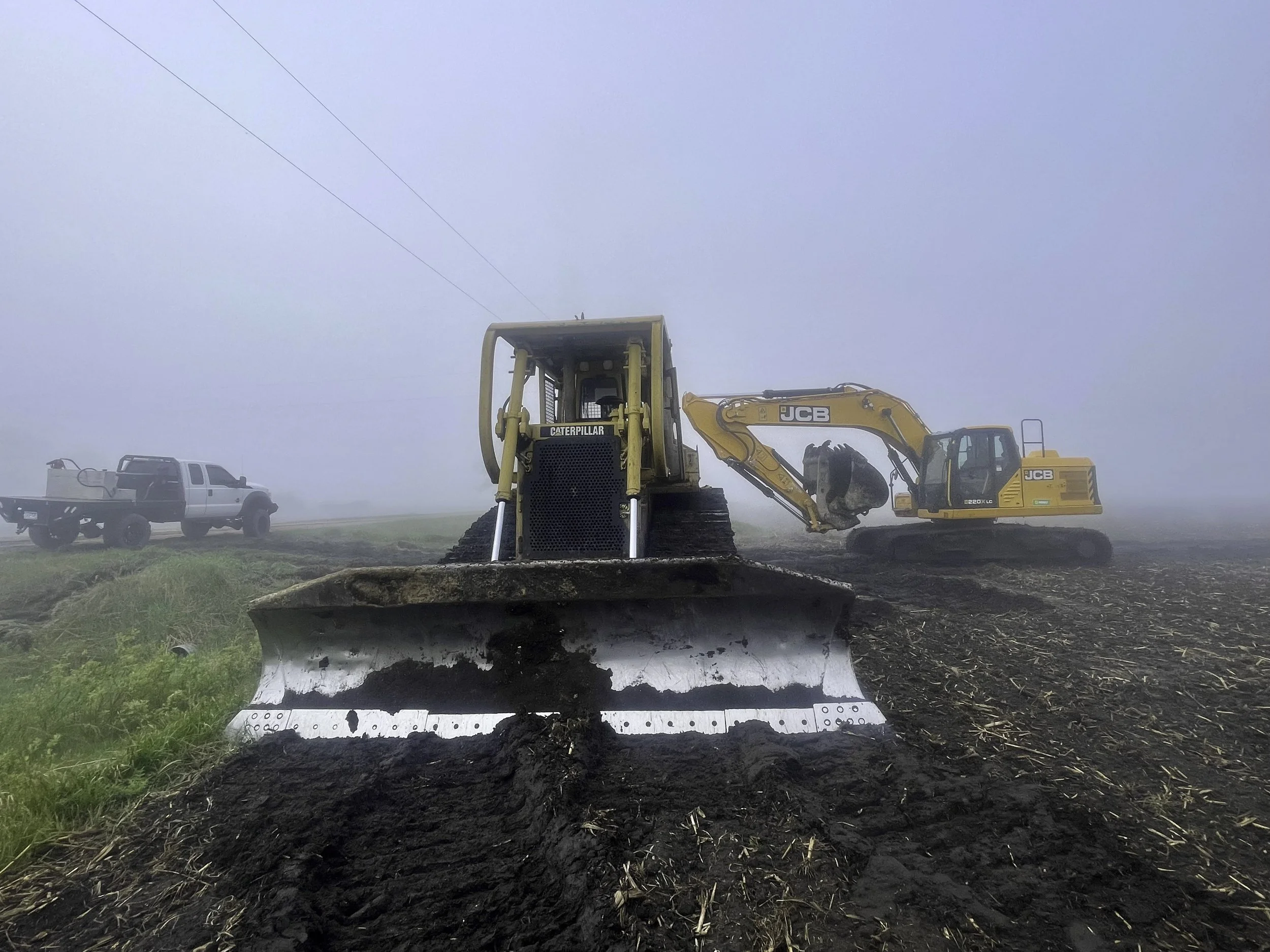 A large bulldozer pushing soil in a foggy field with a JCB excavator and a pickup truck in the background.