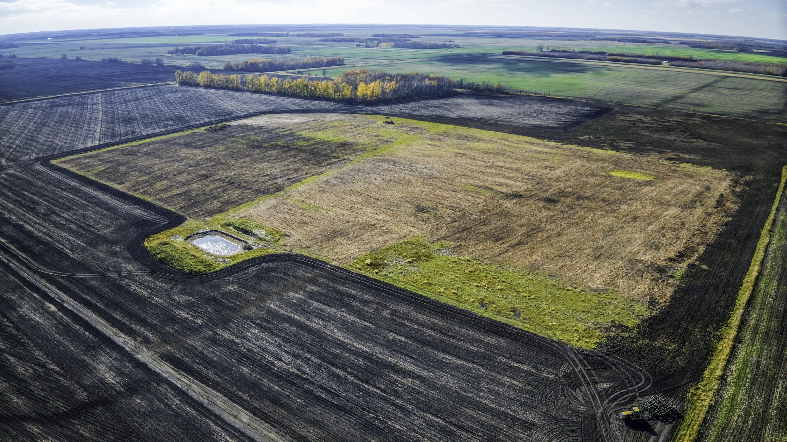 Aerial view of a large agricultural field with different crop colors, a small pond, and farming equipment in the bottom right corner.