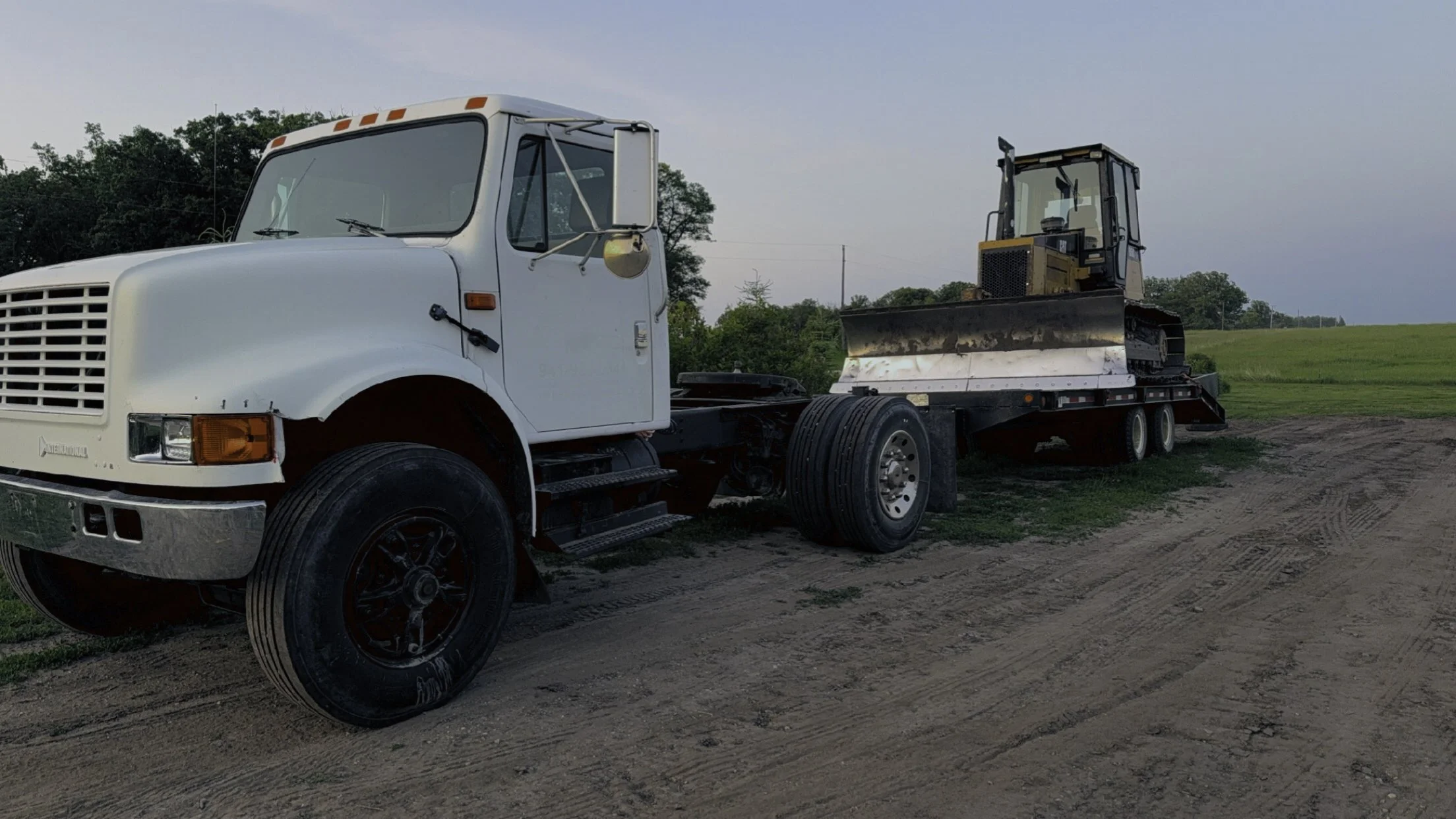 A white flatbed truck with black tires parked on dirt ground, carrying a yellow bulldozer with tracks, in a rural area with green grass and trees in the background.