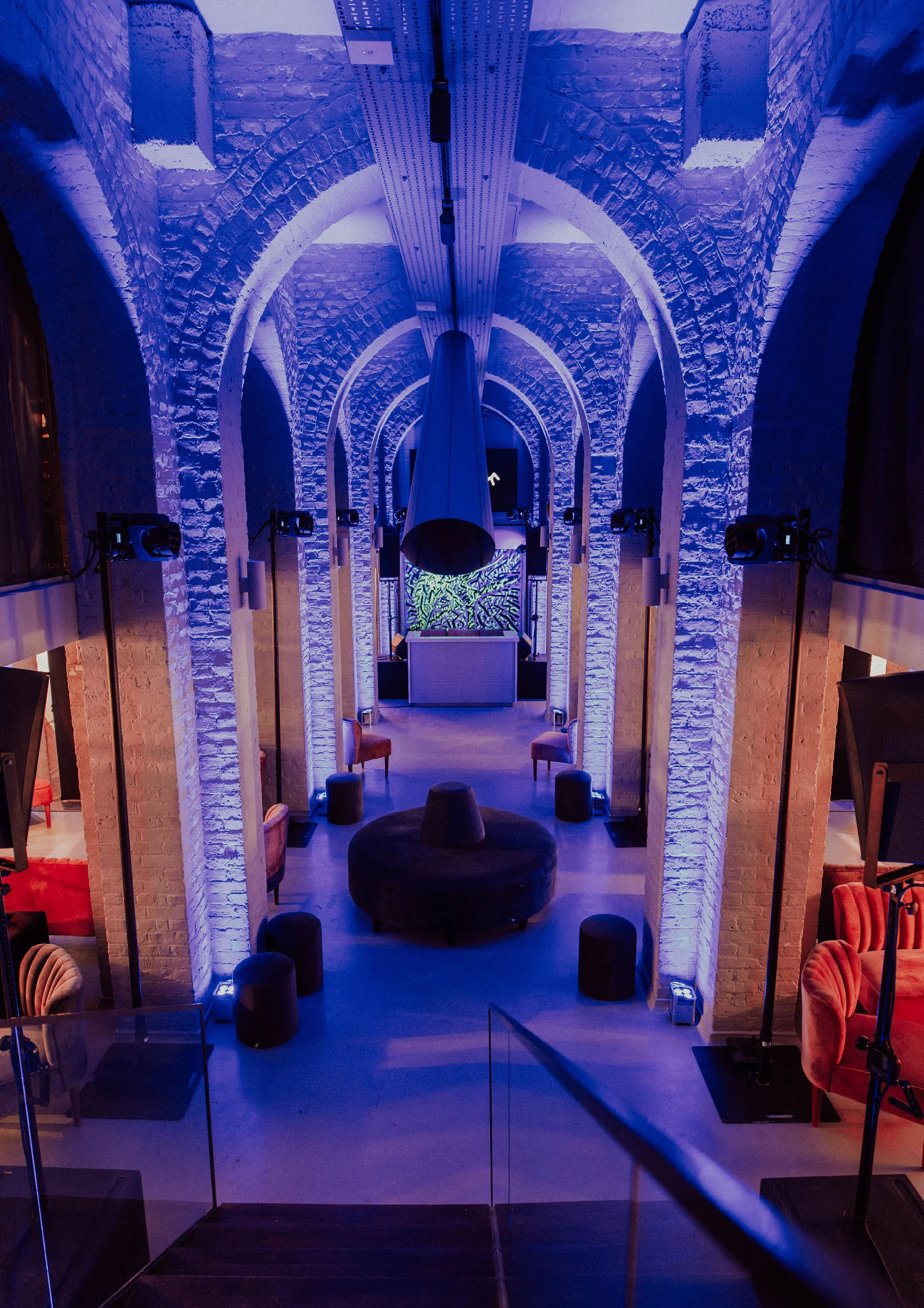Interior of a modern venue with exposed brick walls and arched architectural features illuminated with purple and blue lighting. There are chairs, stools, and a large curved structure in the center, possibly a DJ booth or stage area.