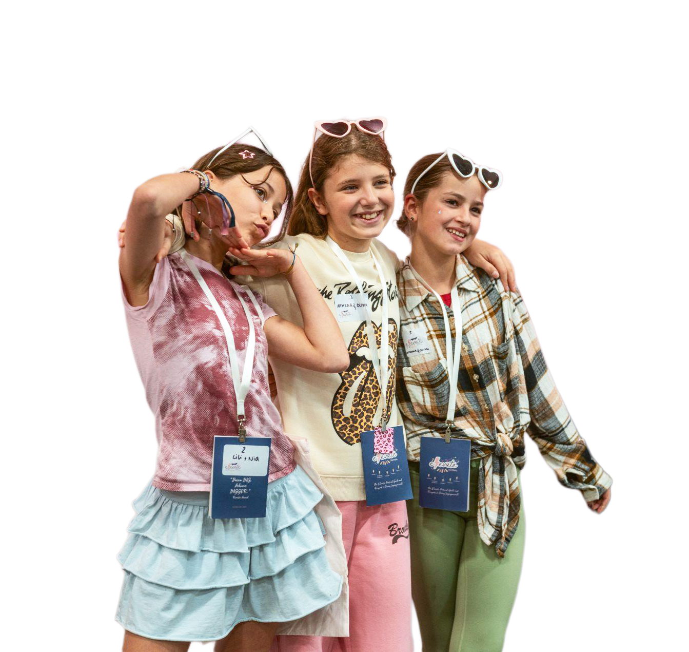 Three young girls standing together, smiling for a photo at an event, wearing casual clothes with sunglasses on their heads and event badges around their necks.
