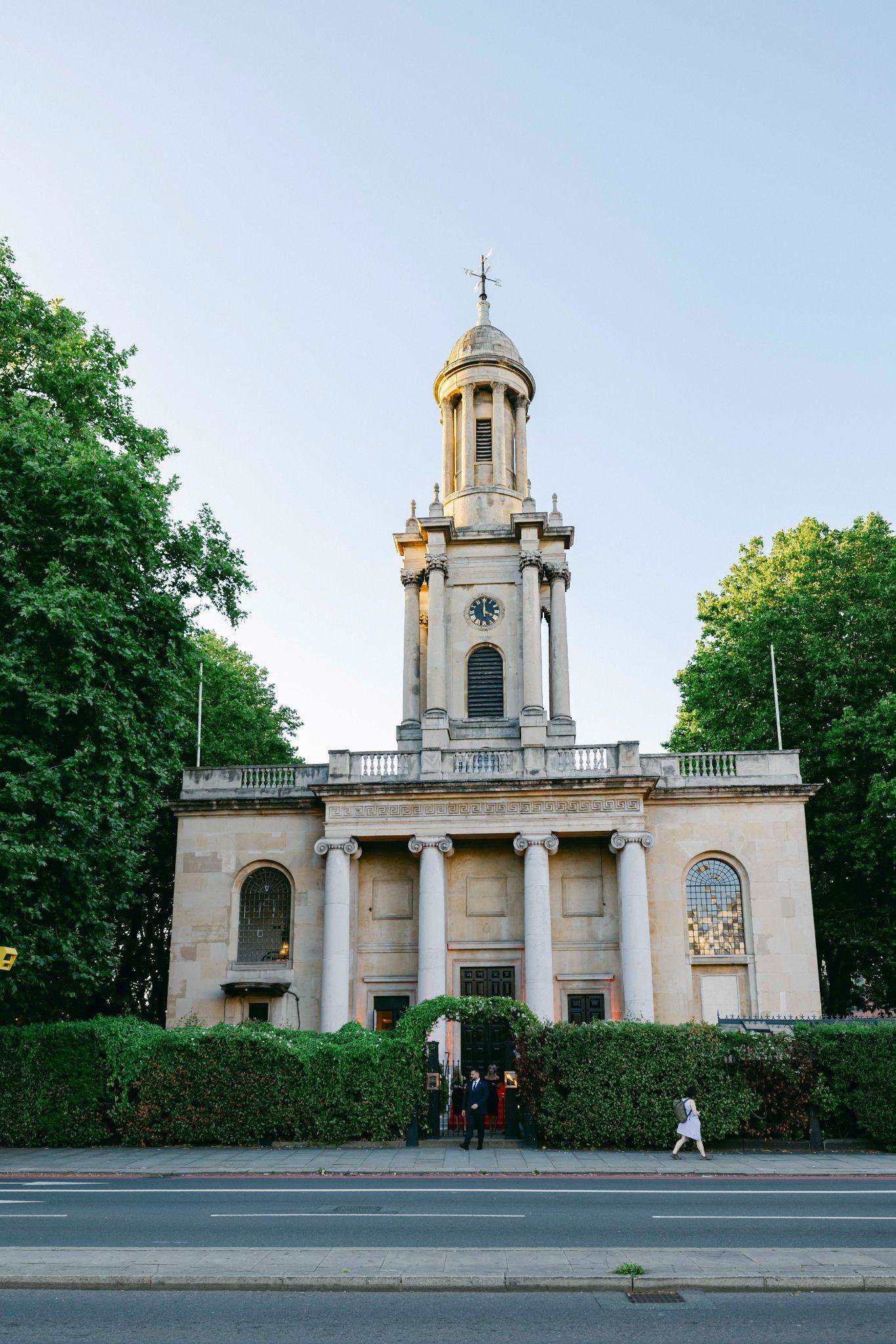 A historic church with a clock tower, stone columns, and an ornate facade, surrounded by green trees and people entering the building.