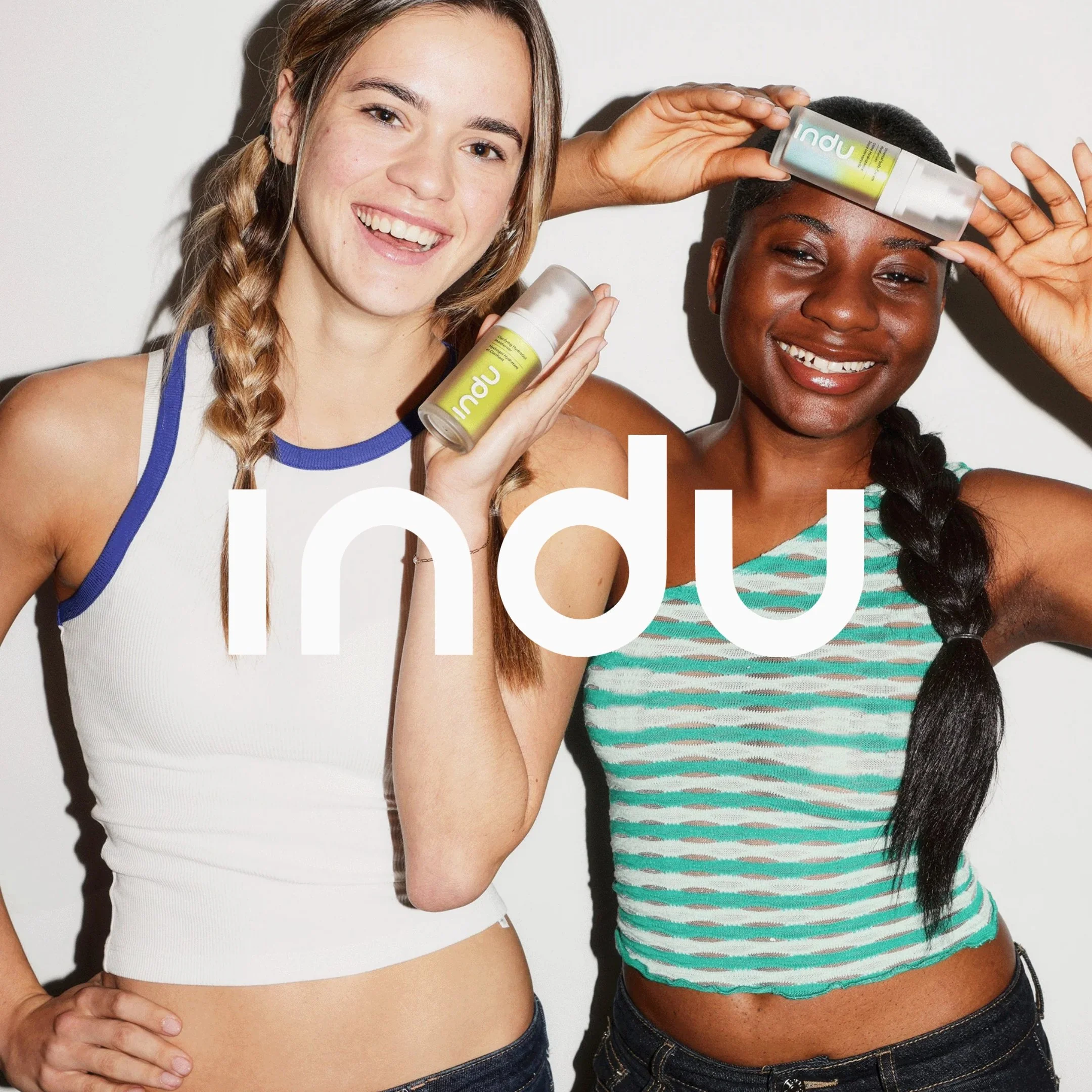 Two women standing side by side smiling, holding skincare products labeled 'Inu,' with one wearing a striped tank top and the other in a white top with blue trim.
