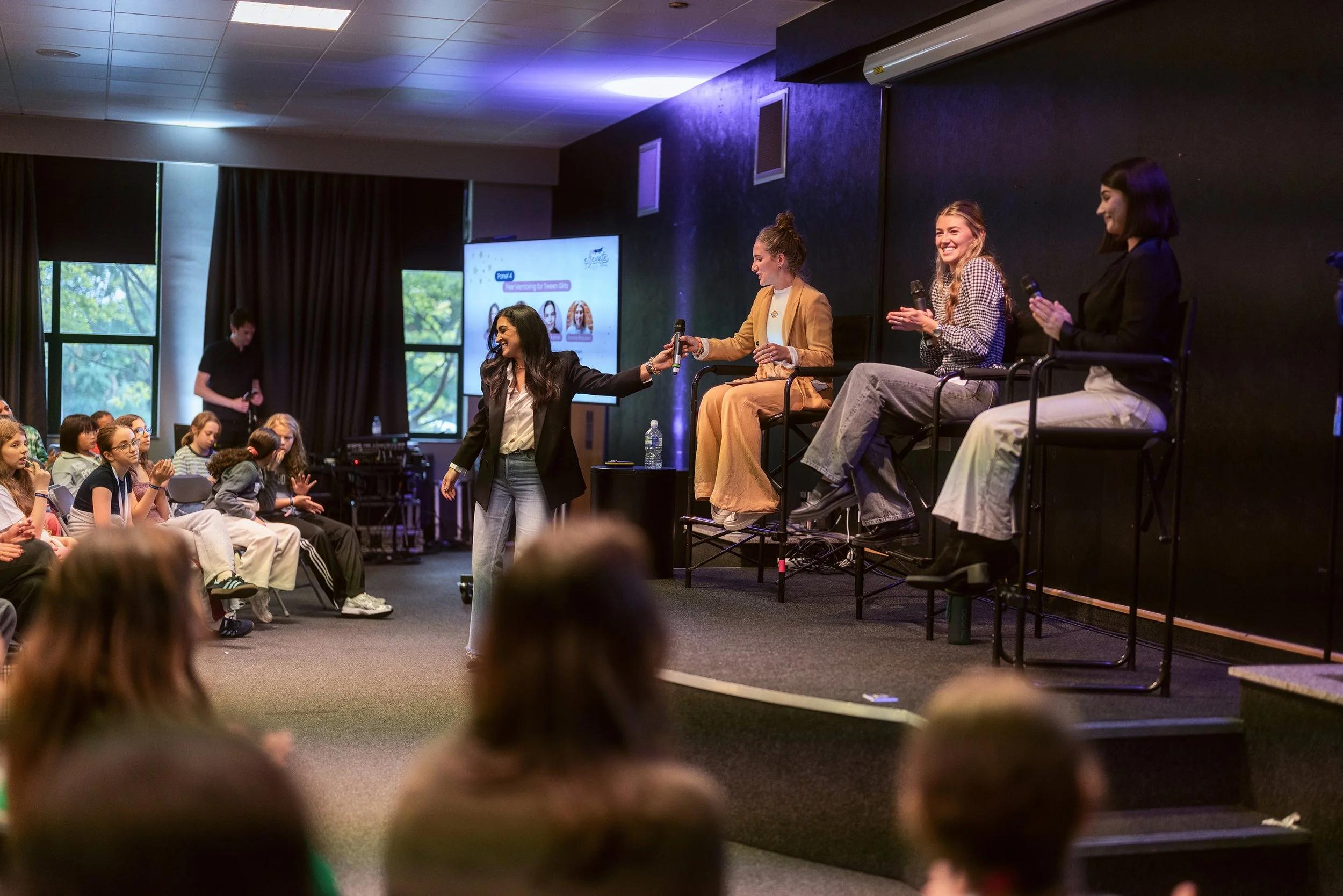A panel discussion at an indoor event with four women seated on stage, while a woman on the stage interacts with the audience. The audience mostly of children, attentively watches and listens. A large screen in the background displays content related to the event.