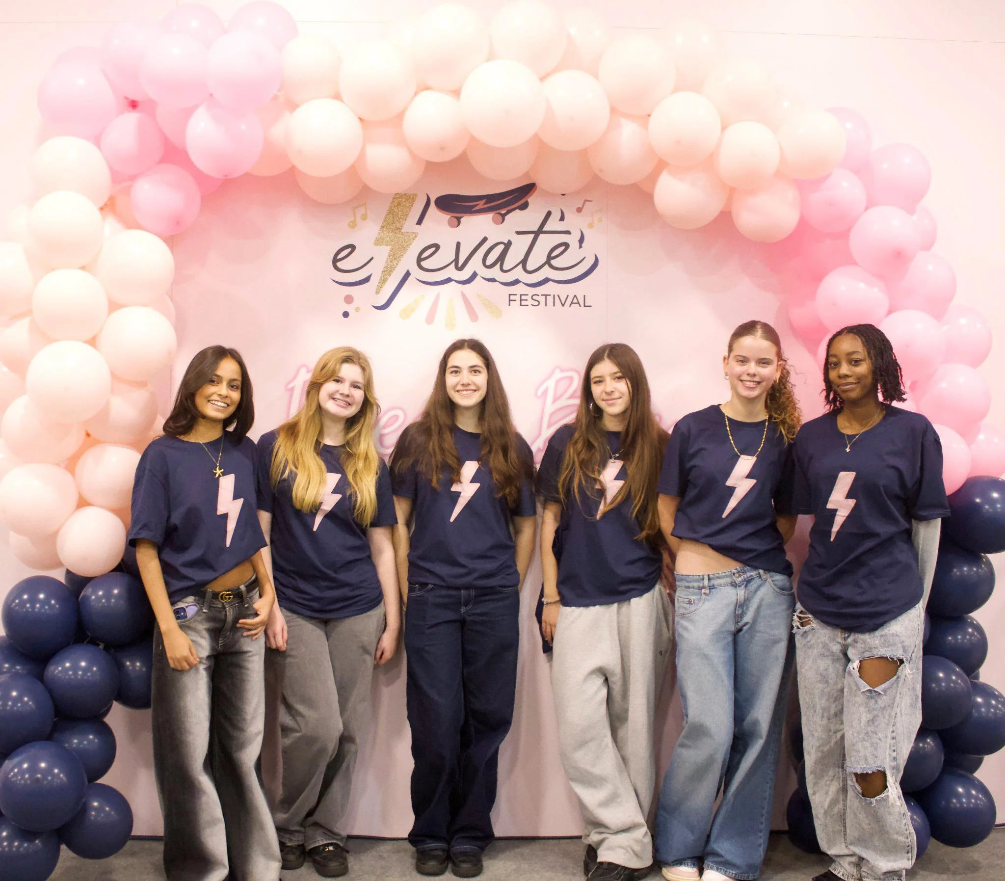Young women standing in front of a pink backdrop decorated with pink and dark blue balloons, with a sign that reads 'Elevate Festival' and features a skateboard and musical notes design.