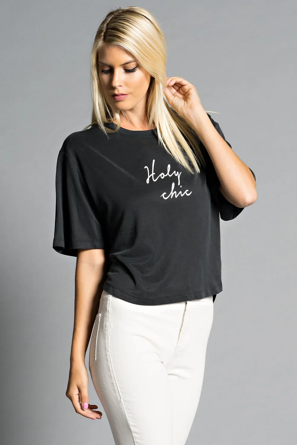 A blonde woman wearing a black T-shirt with white text that says "Holy chic" and white pants, standing against a gray background.