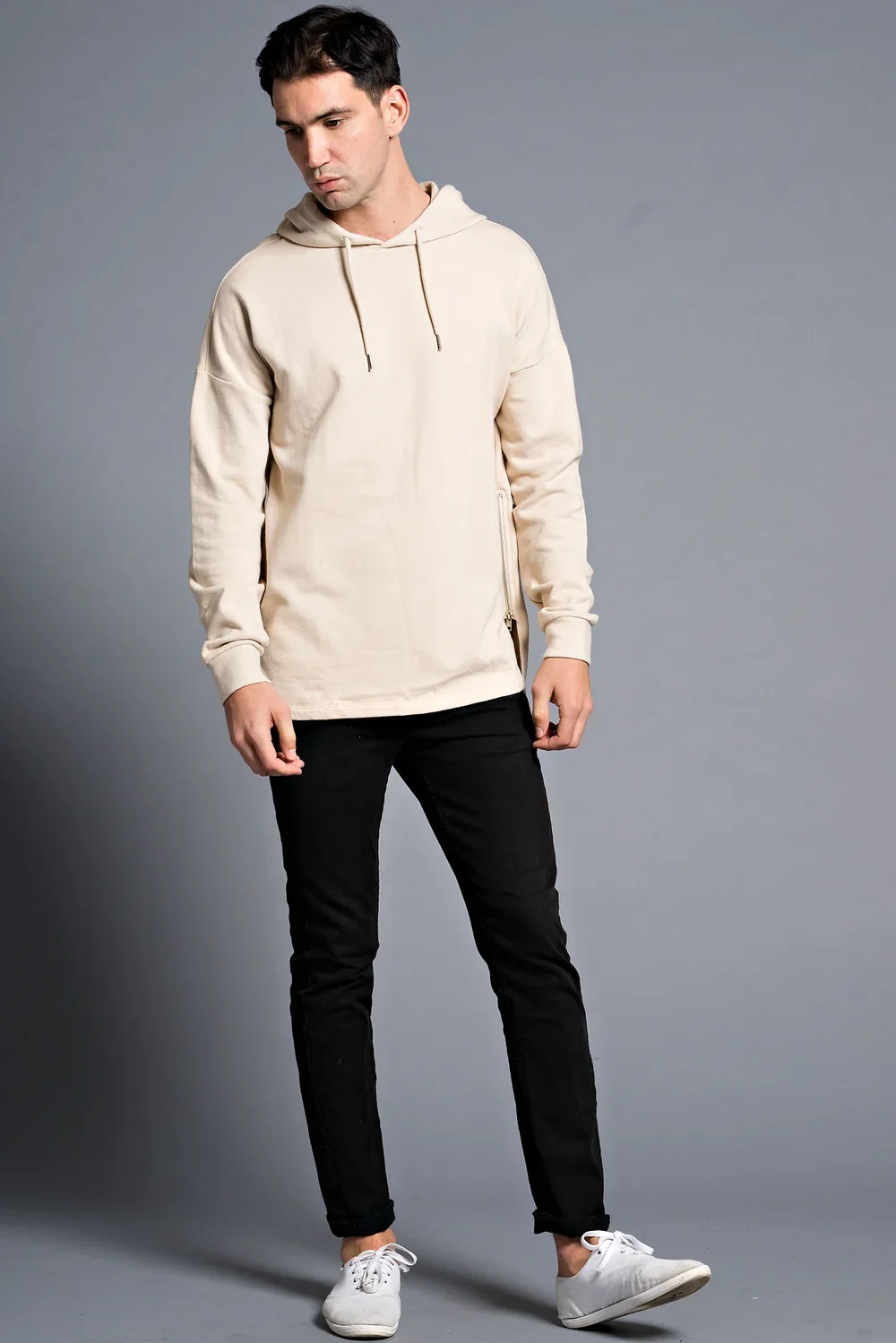 A young man standing against a plain gray background, wearing a beige hoodie, black pants, and white sneakers, looking down slightly.