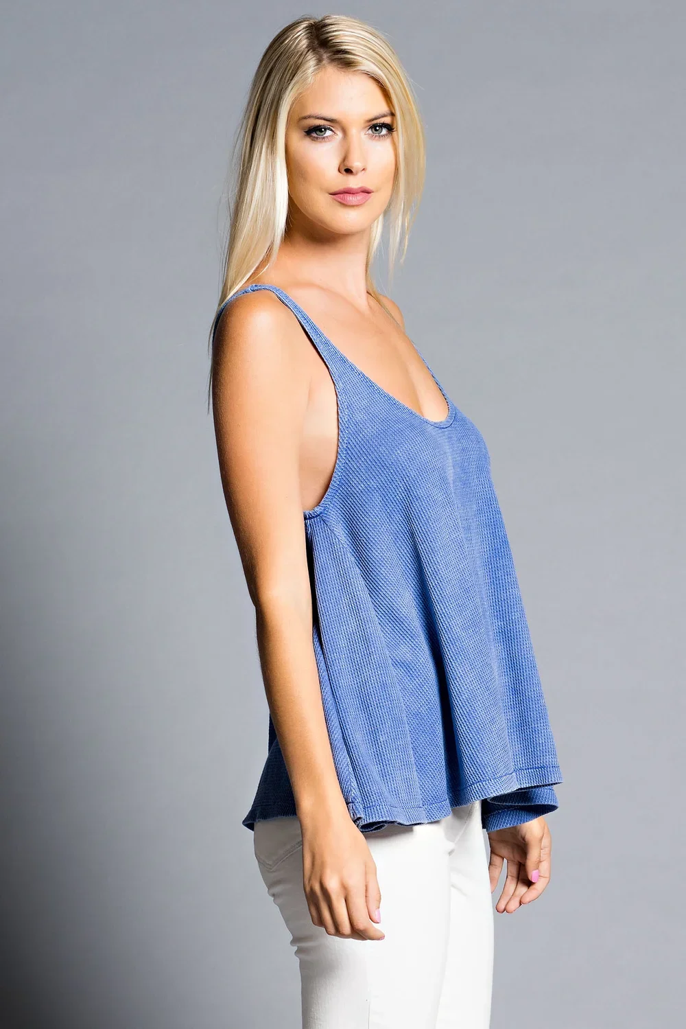 A blonde woman in a blue sleeveless top and white pants standing against a gray background.