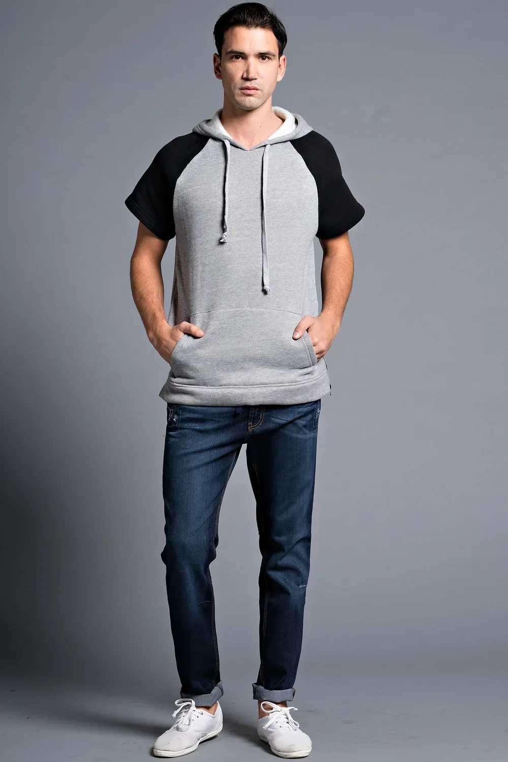 A young man standing against a plain gray background, wearing a short-sleeved hoodie with black and gray sections, blue jeans, and white sneakers, with hands in his hoodie pockets.