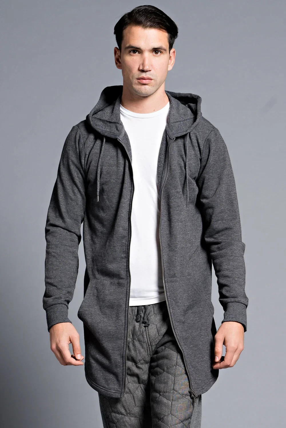 A young man with dark hair and light skin wearing a dark gray zip-up hoodie, white t-shirt, and patterned gray pants, standing against a plain gray background.