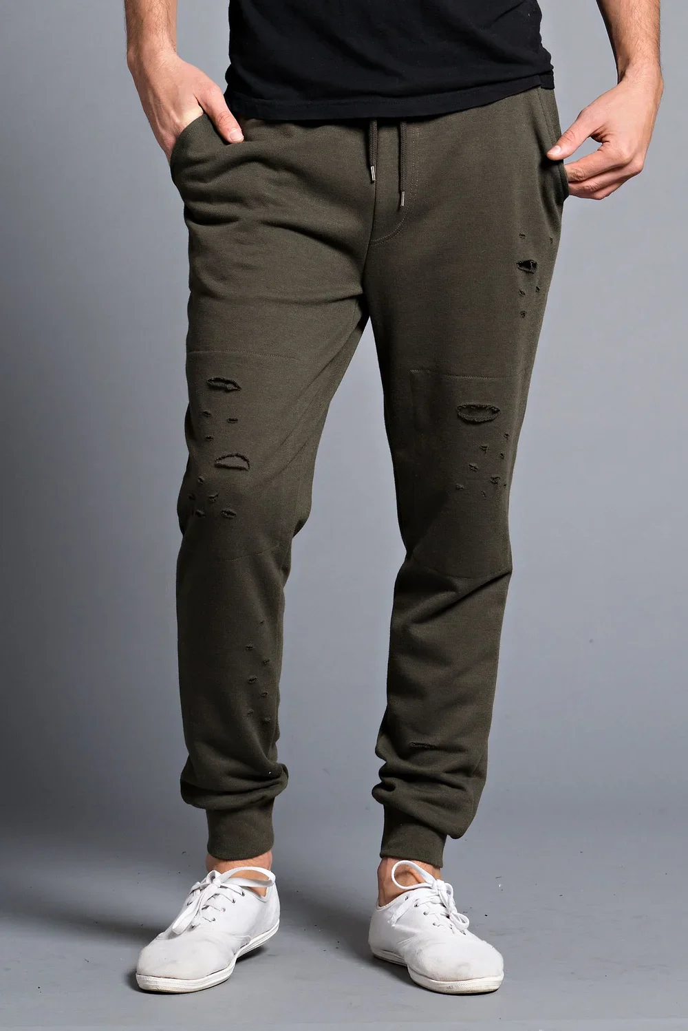 Person wearing distressed olive green sweatpants and white sneakers, standing against a gray background.
