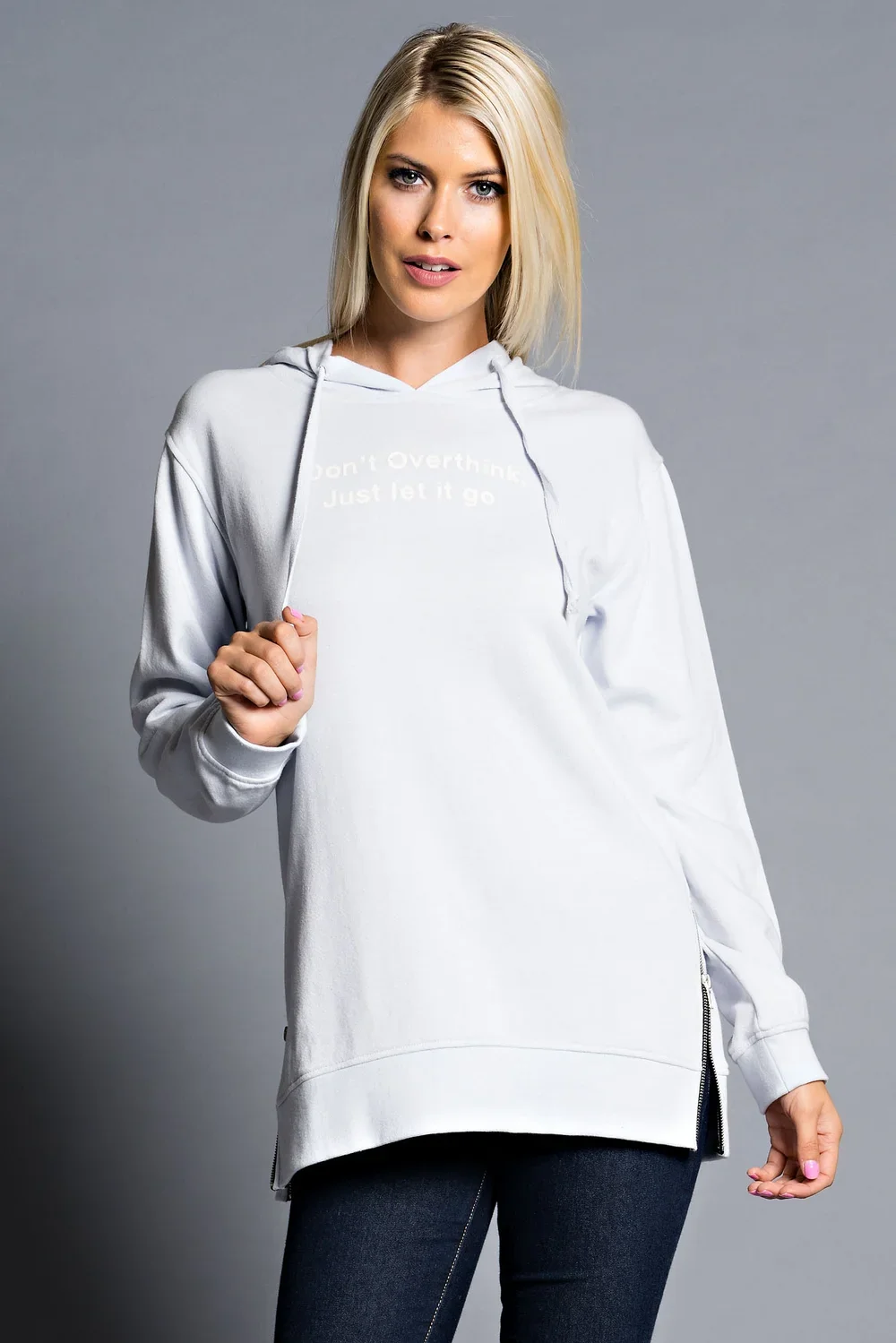 A woman with blonde hair in a white hoodie and dark pants is holding the drawstring of her hoodie with one hand. The hoodie has a slogan that reads, 'Don't Overthink Just let it go.' She is standing against a plain gray background.