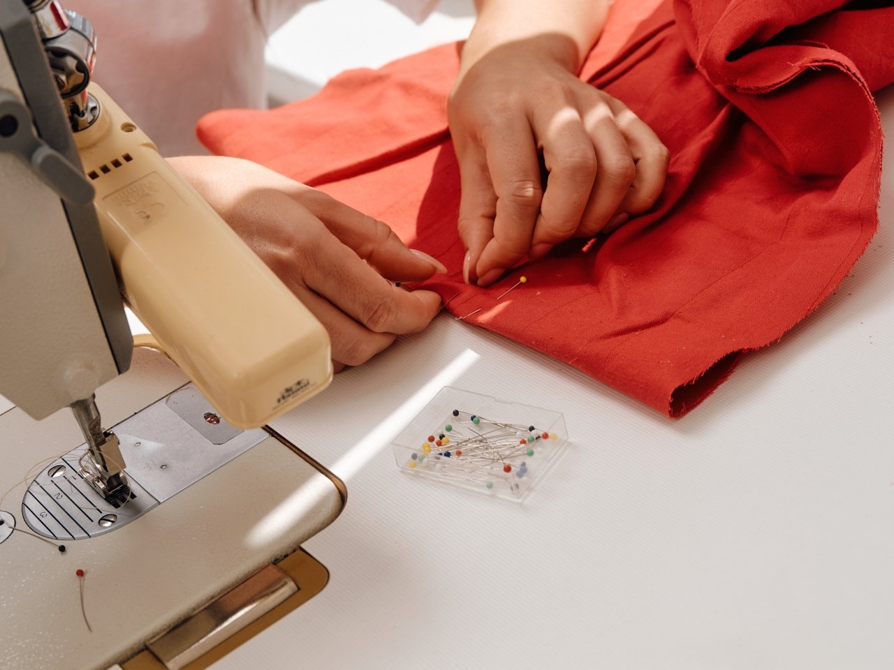 Person sewing red fabric with a sewing machine and a container of pins on the table.