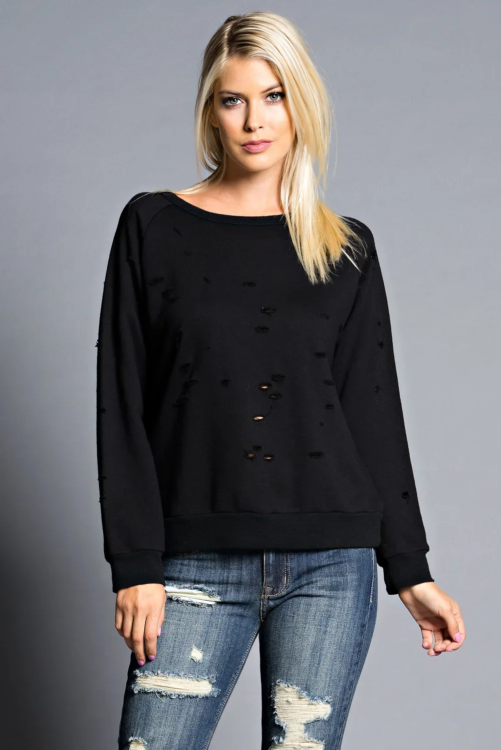 A woman with blonde hair, wearing a black distressed sweatshirt and ripped jeans, stands against a gray background.